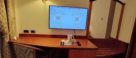 A hotel room with a desk, a chair, a lamp, and a digital screen displaying hotel information.