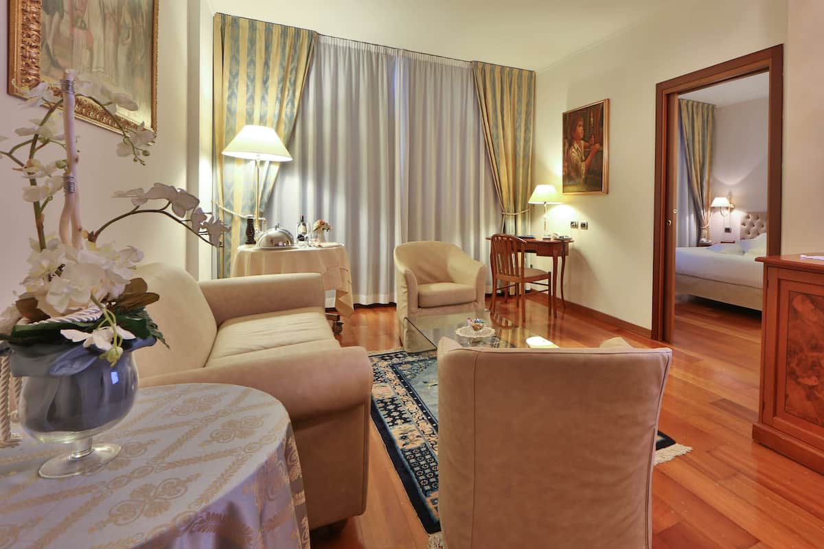 A hotel room with a sofa, armchair, small round table, and a glass coffee table. There is a bed visible through an open door, a painting on the wall, and a window with curtains.