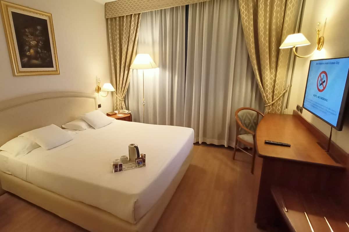 A hotel room with a bed, bedside table, lamp, chair, desk, and a television displaying a no-smoking sign.