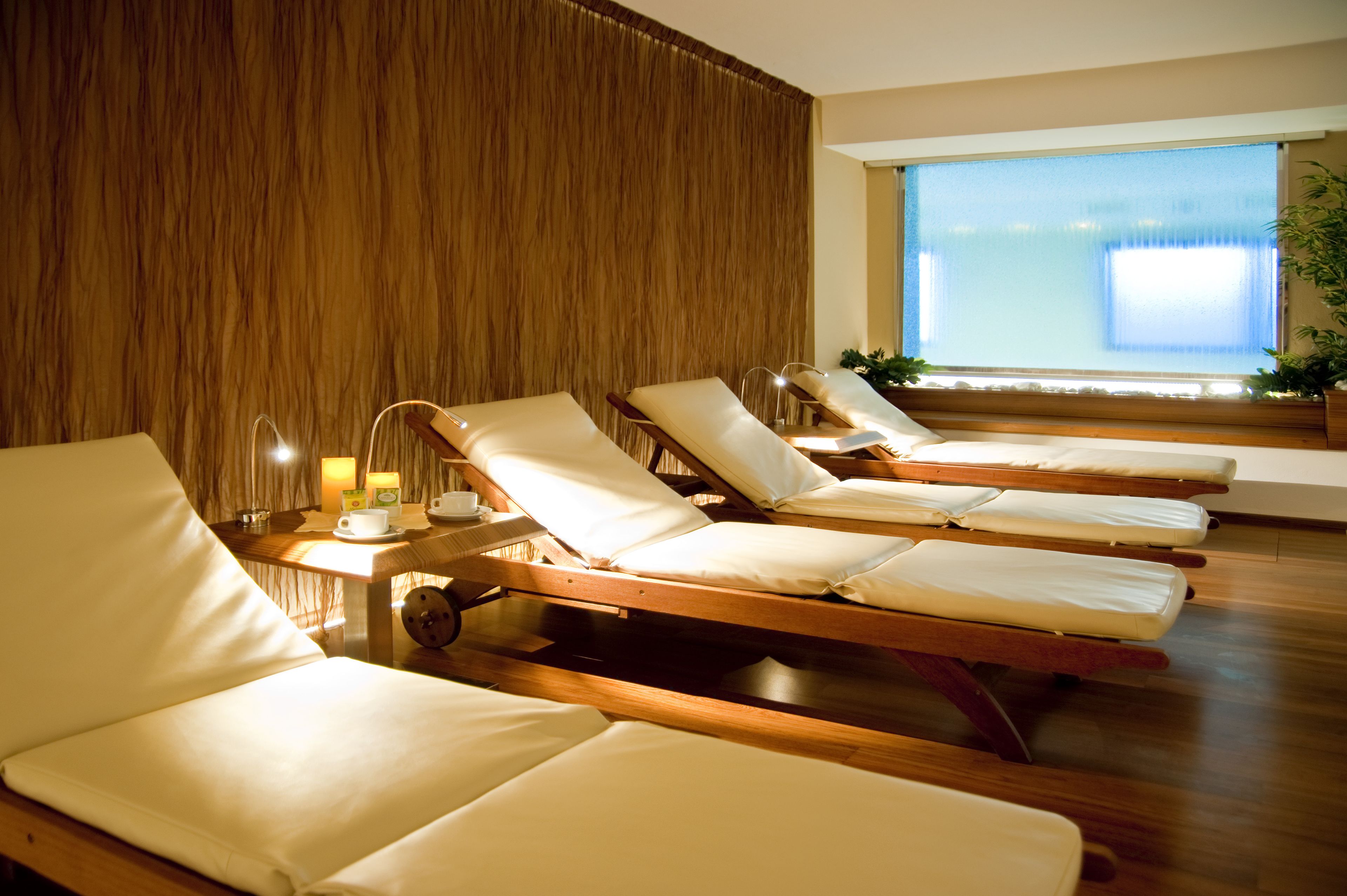 A room with four sunbeds, a wooden wall, a window with a view, and a small table with a lamp and a cup.