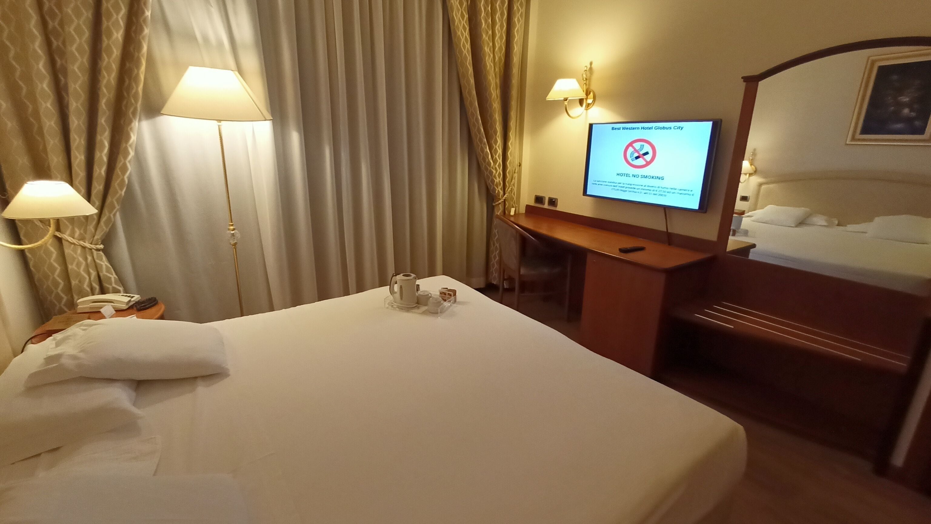 A hotel room with a bed, a desk, a chair, a TV displaying a no-smoking sign, and a lamp.