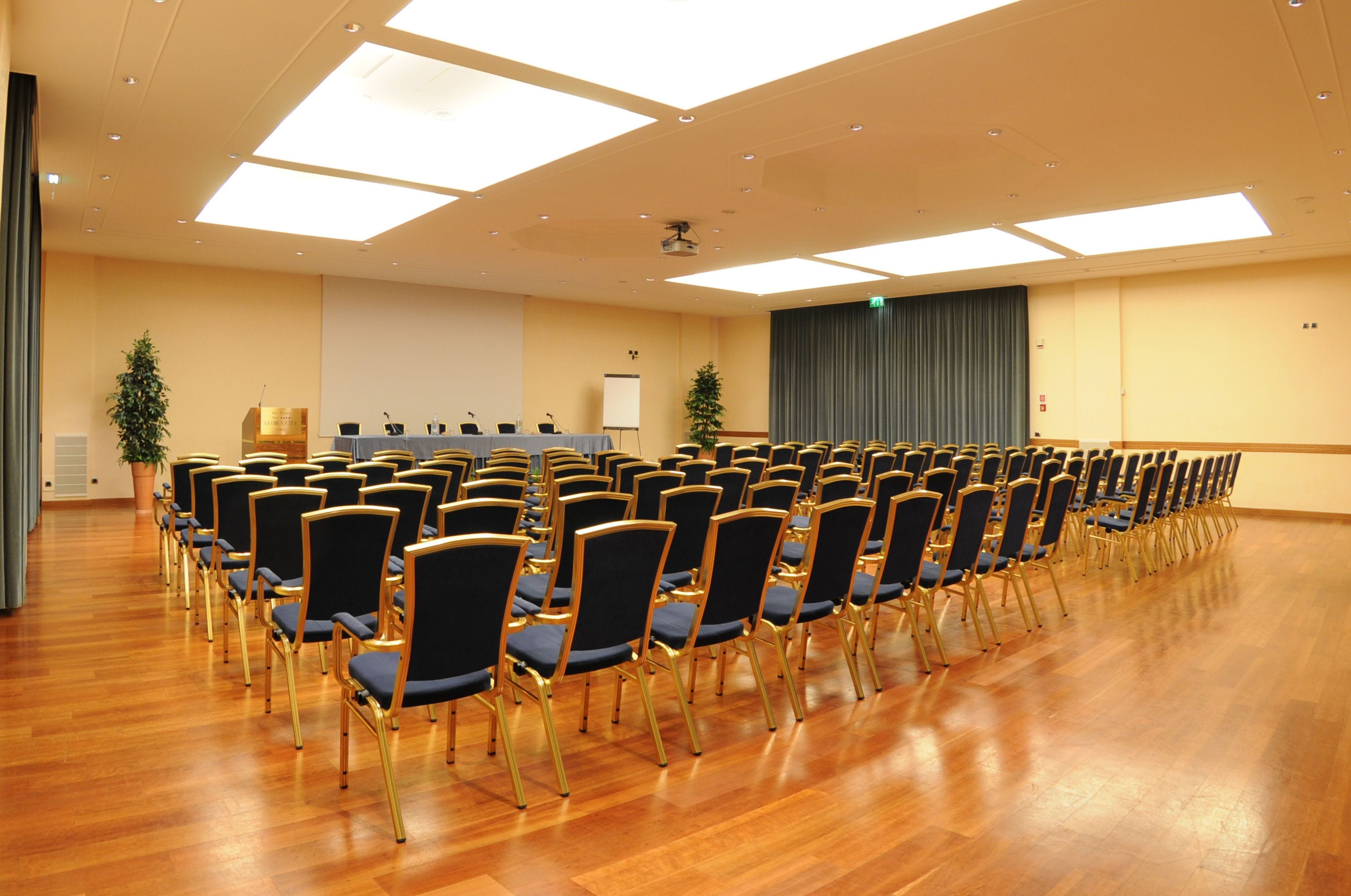 Un salón de conferencias con filas de sillas, un atril y una pantalla para proyector.