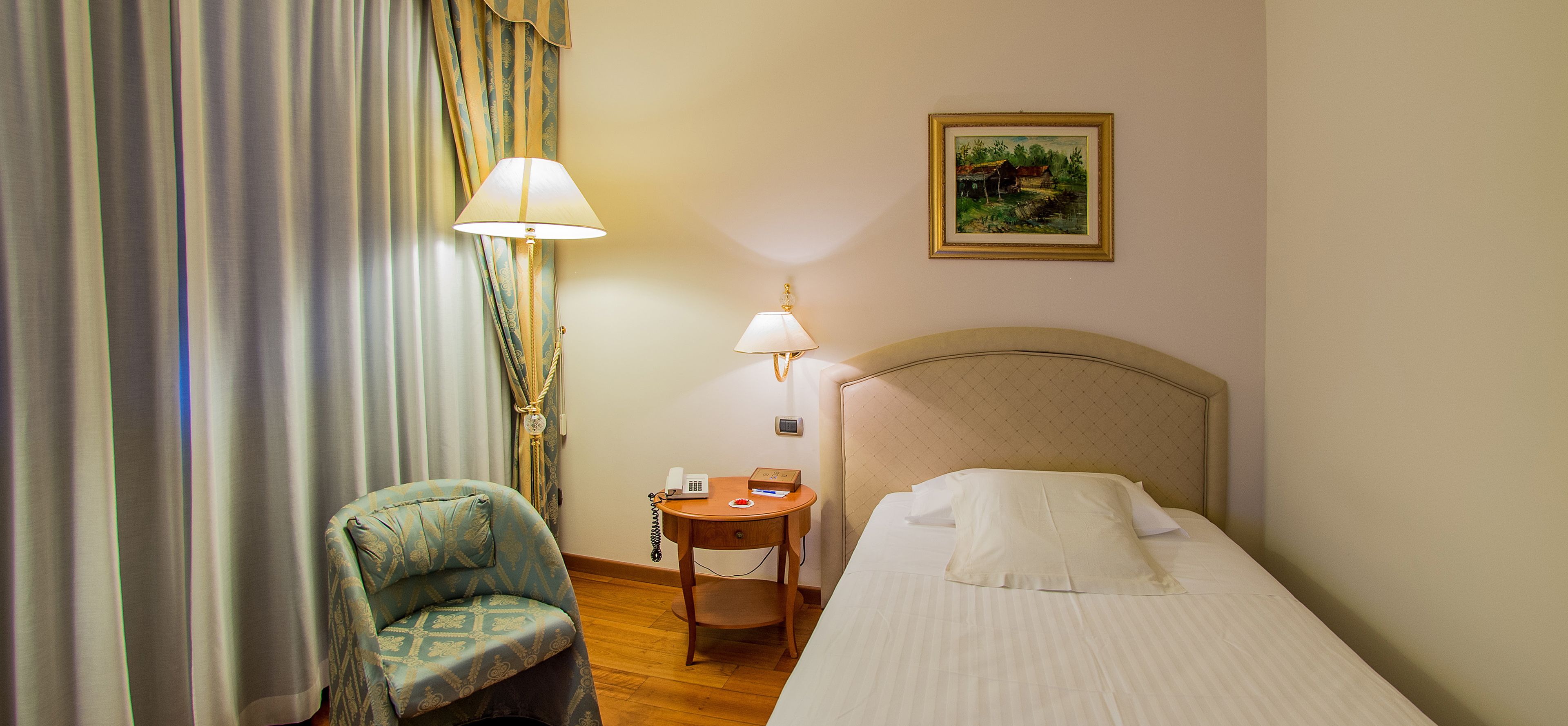A hotel room with a bed, a chair, a nightstand with a lamp, and a painting on the wall.