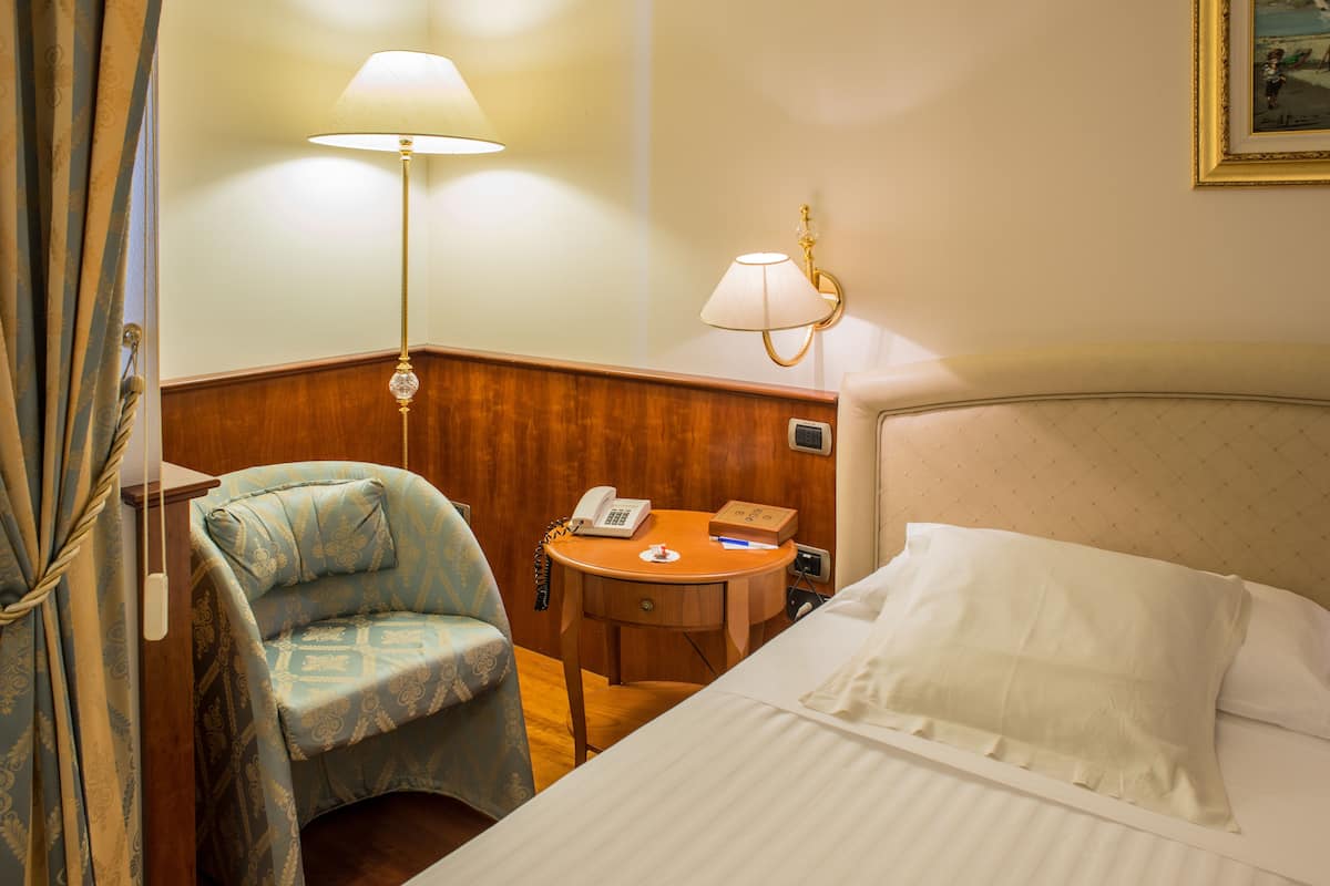 A hotel room with a bed, a desk with a telephone, a chair, and two wall-mounted lamps.