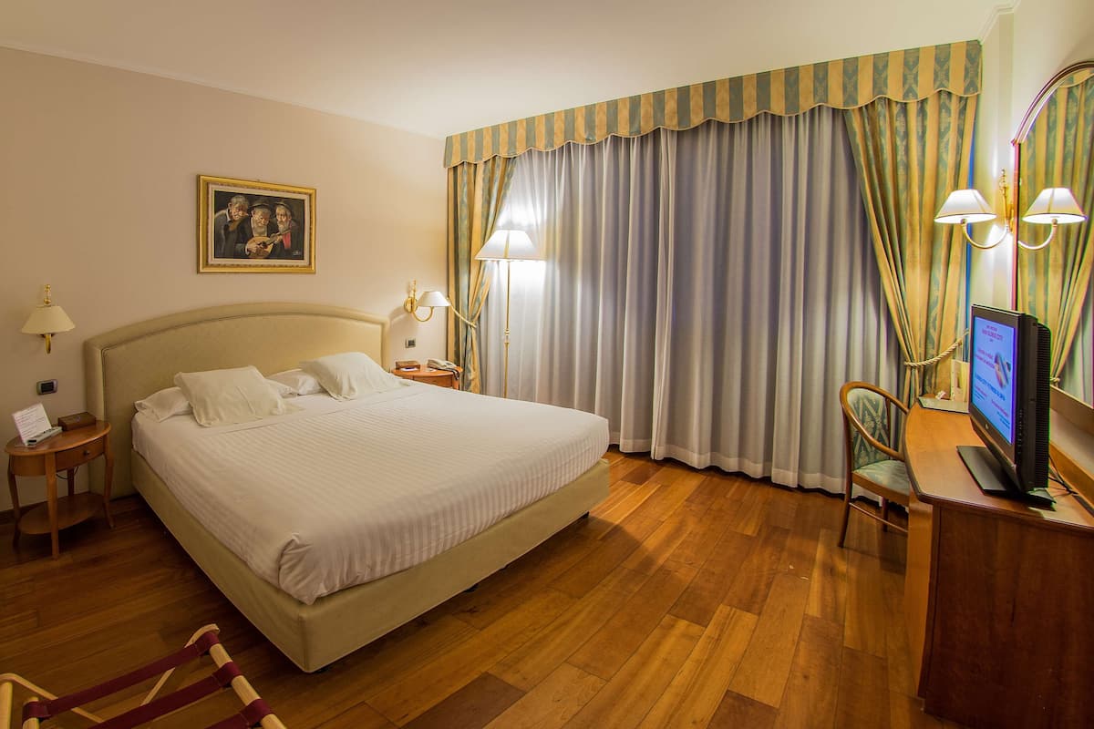 A hotel room with a large bed, wooden flooring, a desk with a computer, a chair, a lamp, and curtains.
