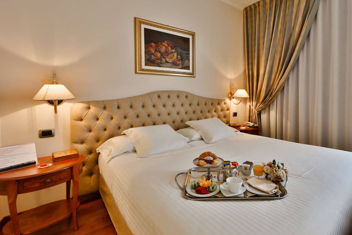 A hotel room with a tufted headboard, a bed with white linens, a tray with breakfast items, a desk with a lamp, and a painting on the wall.