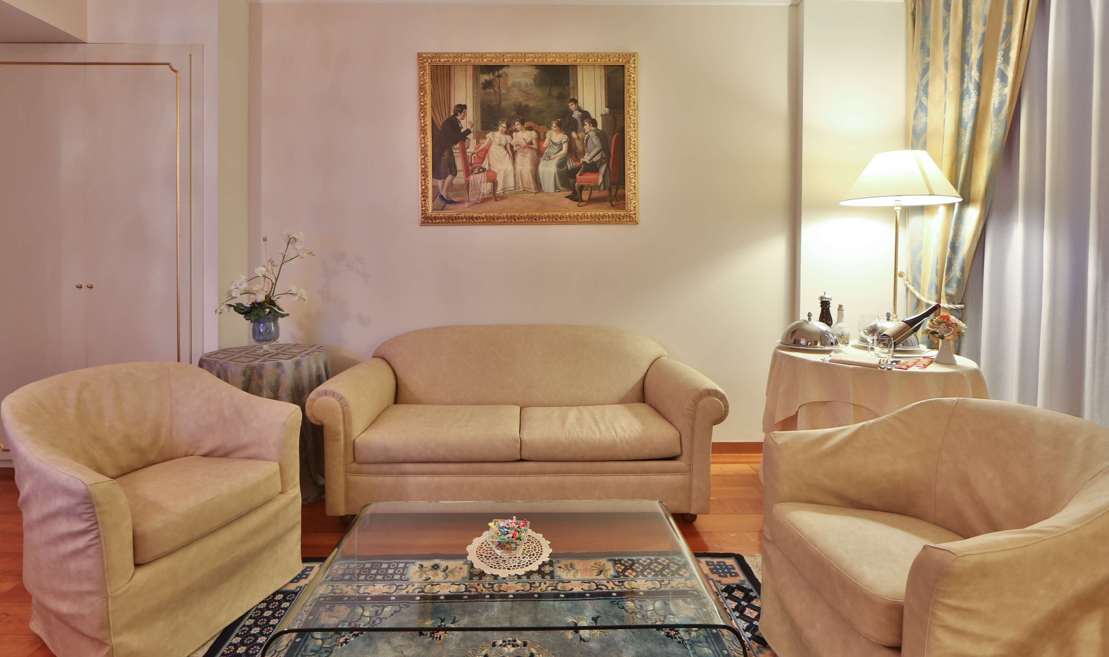 A living room with a beige sofa, two armchairs, a glass coffee table, a painting on the wall, a lamp, and a small table with a vase and a bottle.