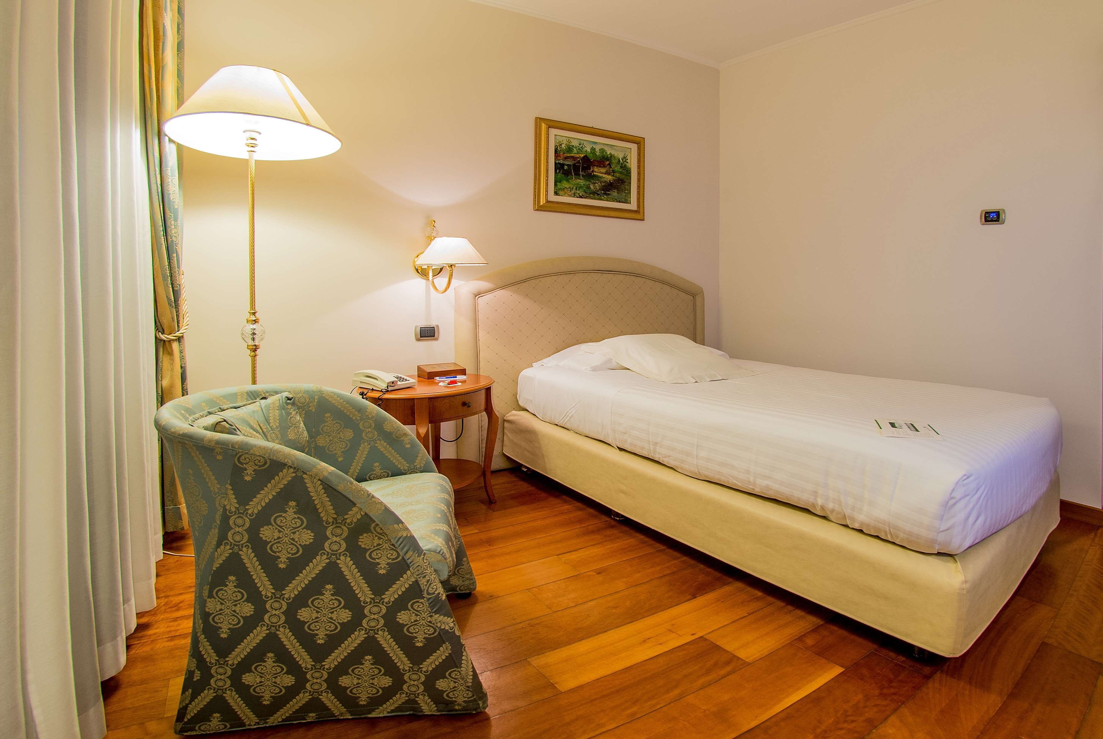 Ein Hotelzimmer mit Bett, Sessel, Lampe und einem Gemälde an der Wand.