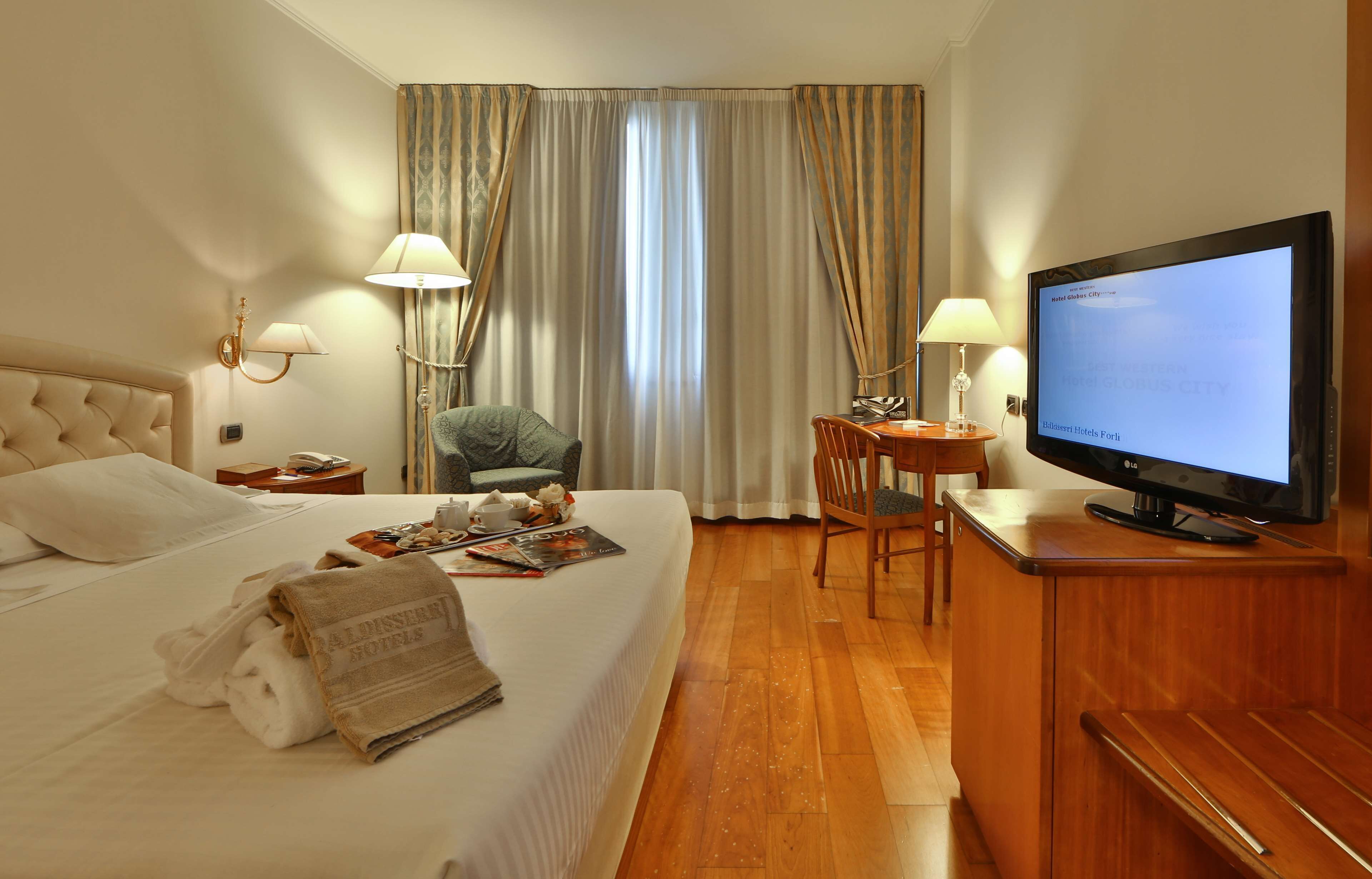 A hotel room with a bed, a desk with a television, a chair, and a lamp.