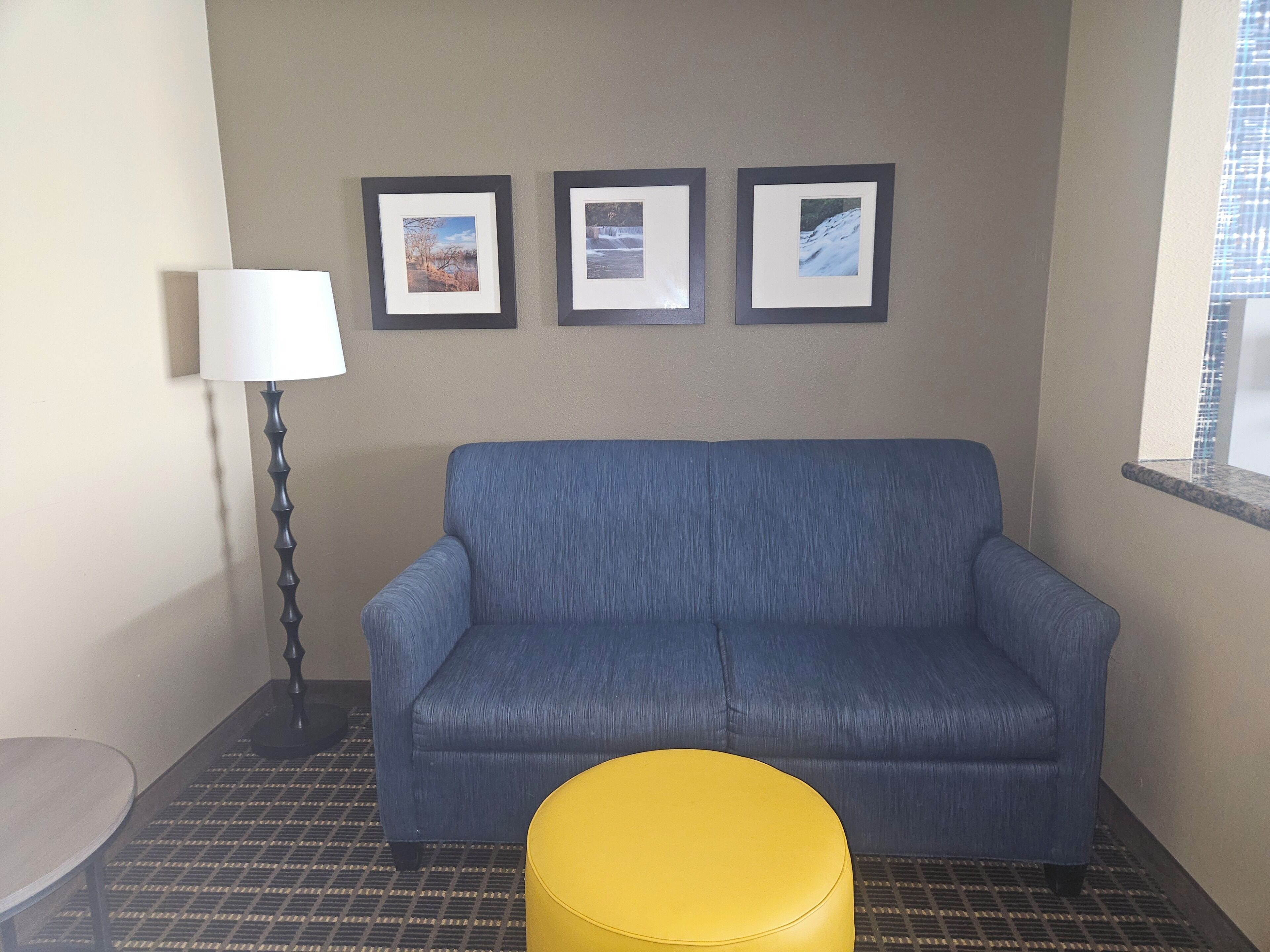 A room with a blue sofa, a yellow ottoman, a lamp, and three framed pictures on the wall.