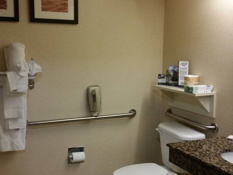 A bathroom with a toilet, handrails, a towel dispenser, a fire alarm, and a sink with a countertop.