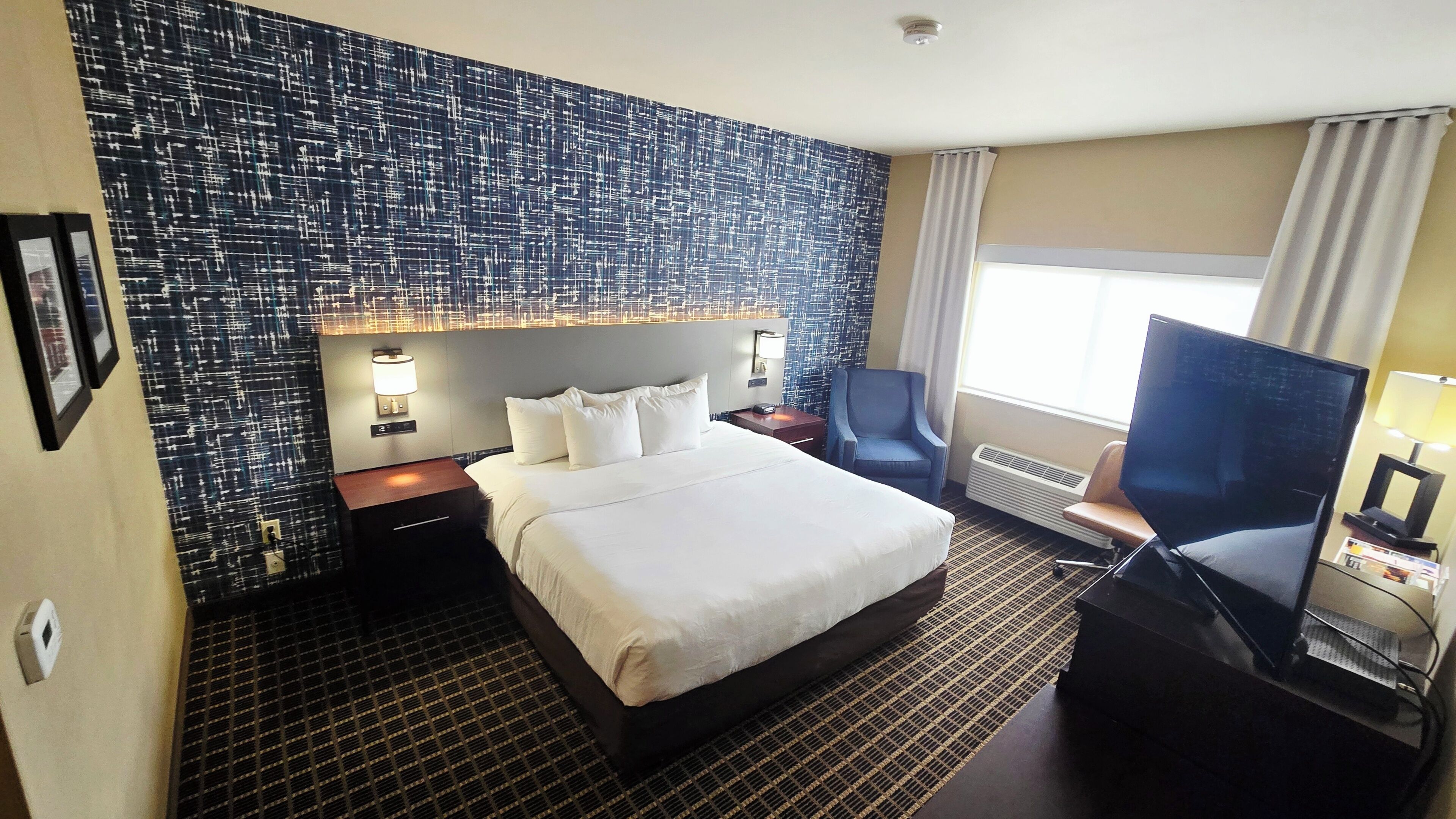 A hotel room with a large bed, a desk, a chair, a TV, and a patterned wall.