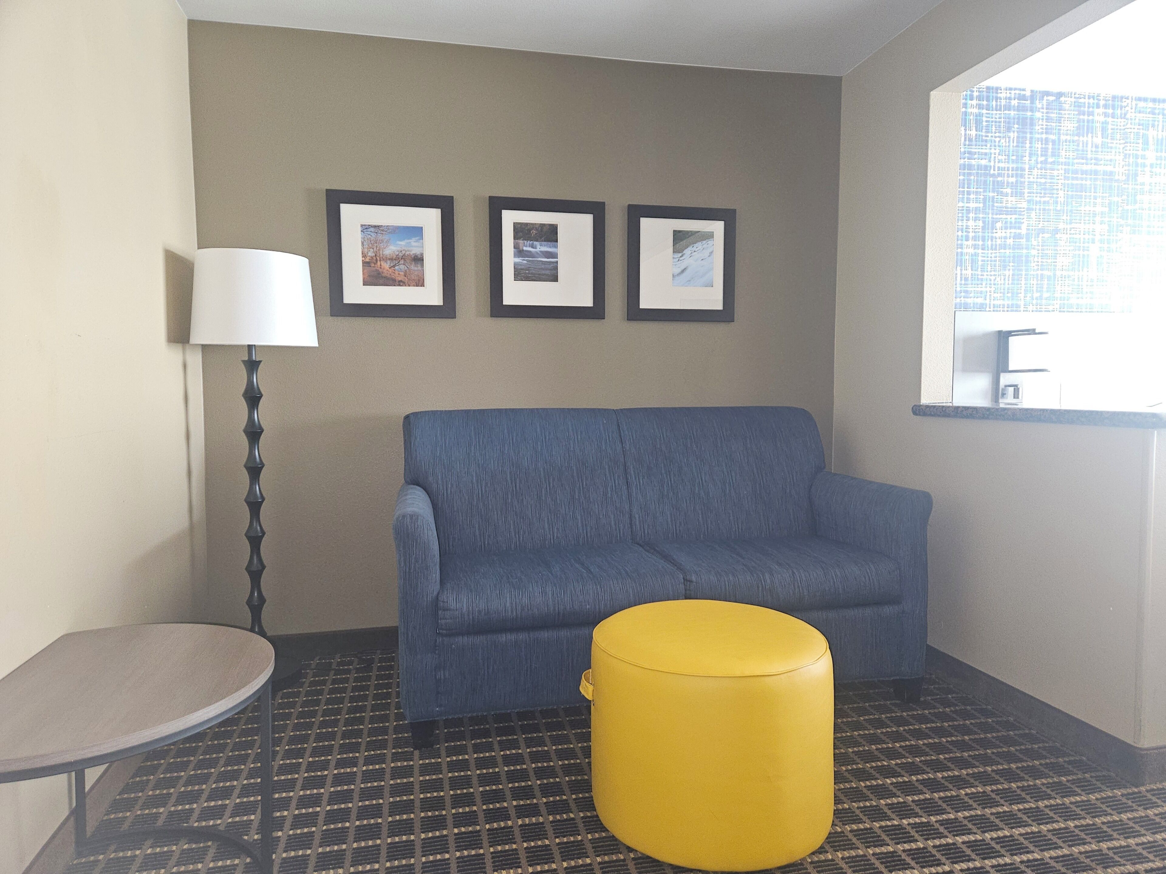 A room with a blue sofa, a yellow ottoman, a small table, and a lamp. There are three framed pictures on the wall.