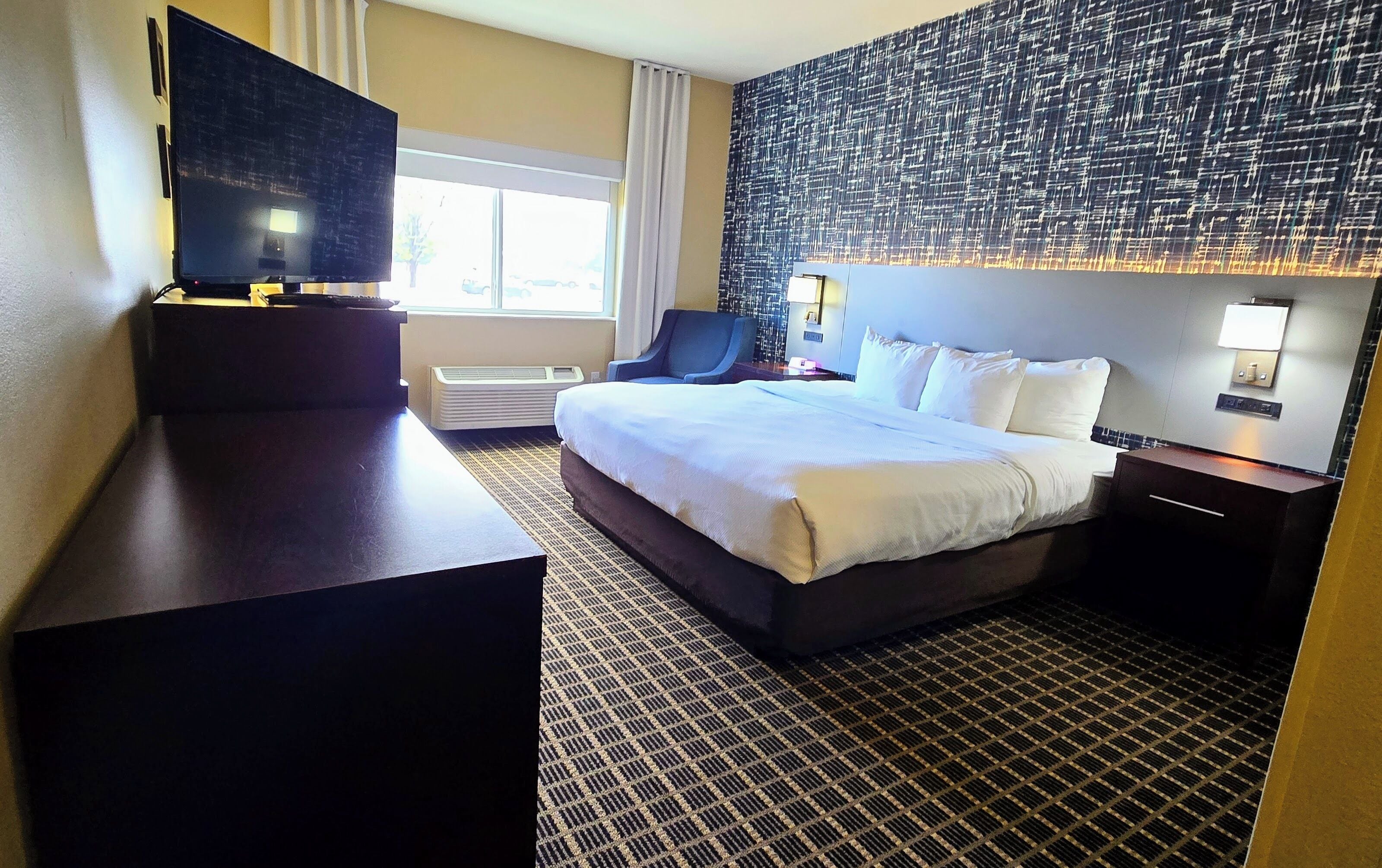A hotel room with a large bed, a desk, a chair, a flat-screen TV, and a patterned wall.