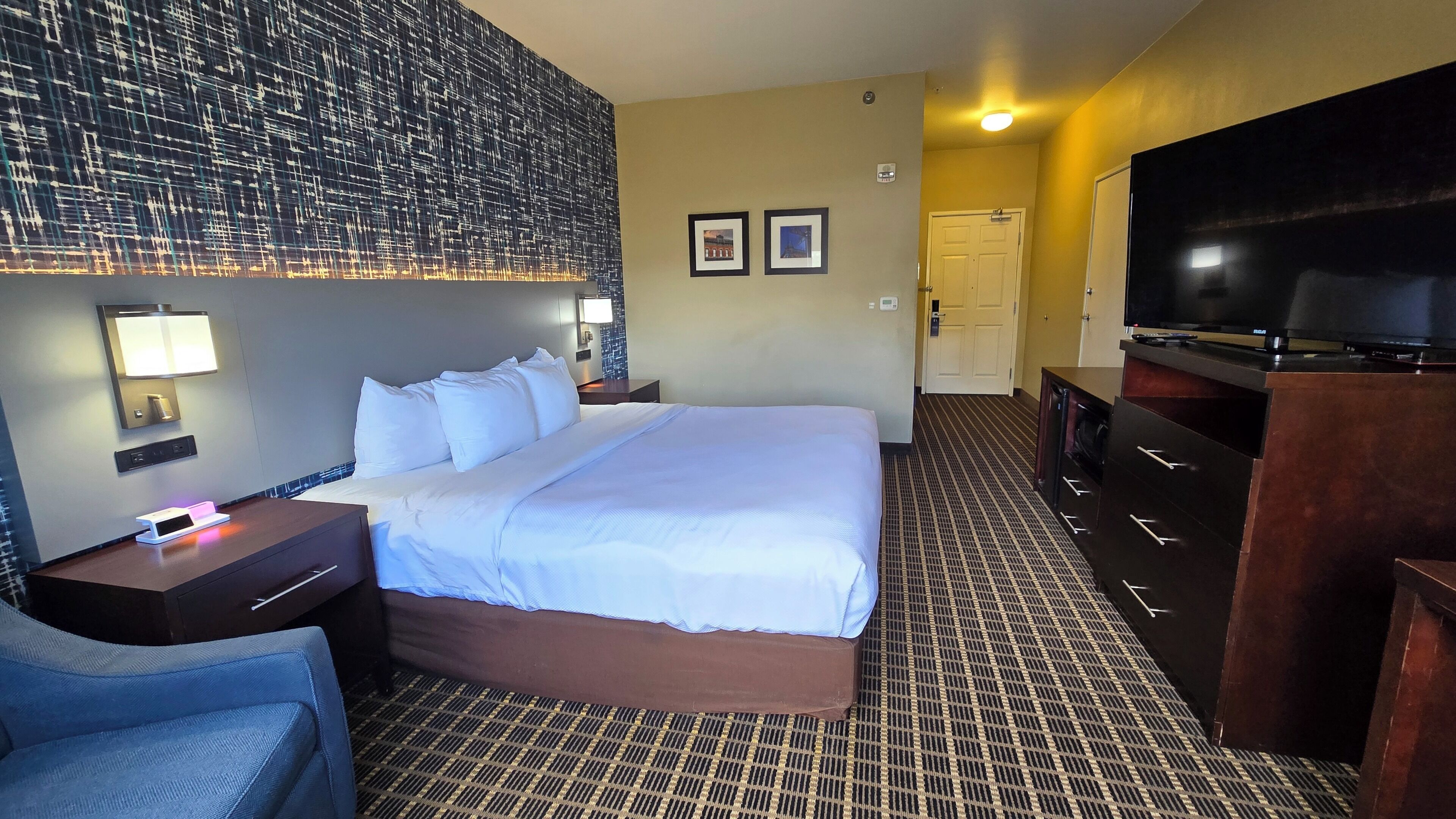Habitación de hotel con una cama grande, un escritorio con lámpara, un televisor y una lámpara de pared.