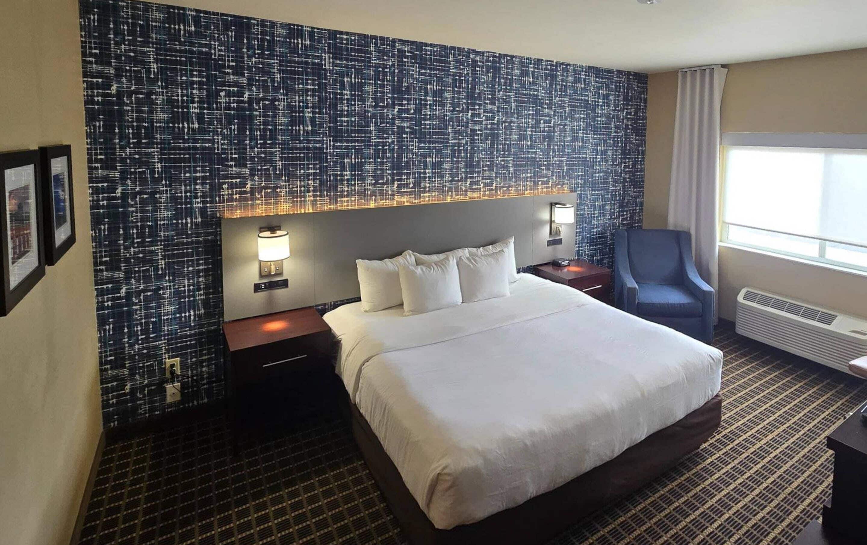 Ein Hotelzimmer mit einem großen Bett, zwei Nachttischen, einem Sessel und einer Wand mit strukturierter Wandgestaltung sowie einem Fenster mit Vorhängen.