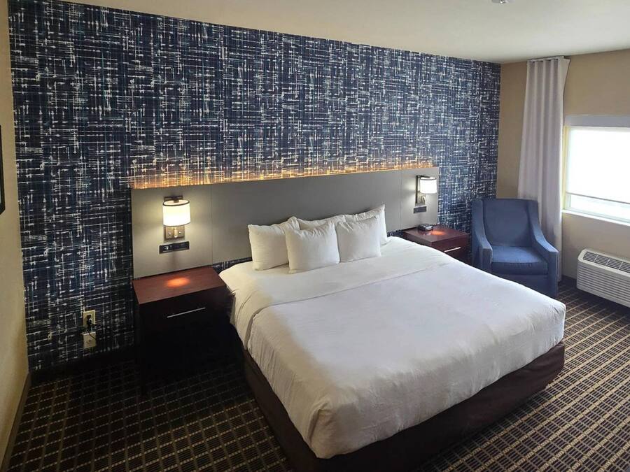 Ein Hotelzimmer mit einem großen Bett, zwei Nachttischen, einem Sessel und einer Wand mit strukturierter Wandgestaltung sowie einem Fenster mit Vorhängen.
