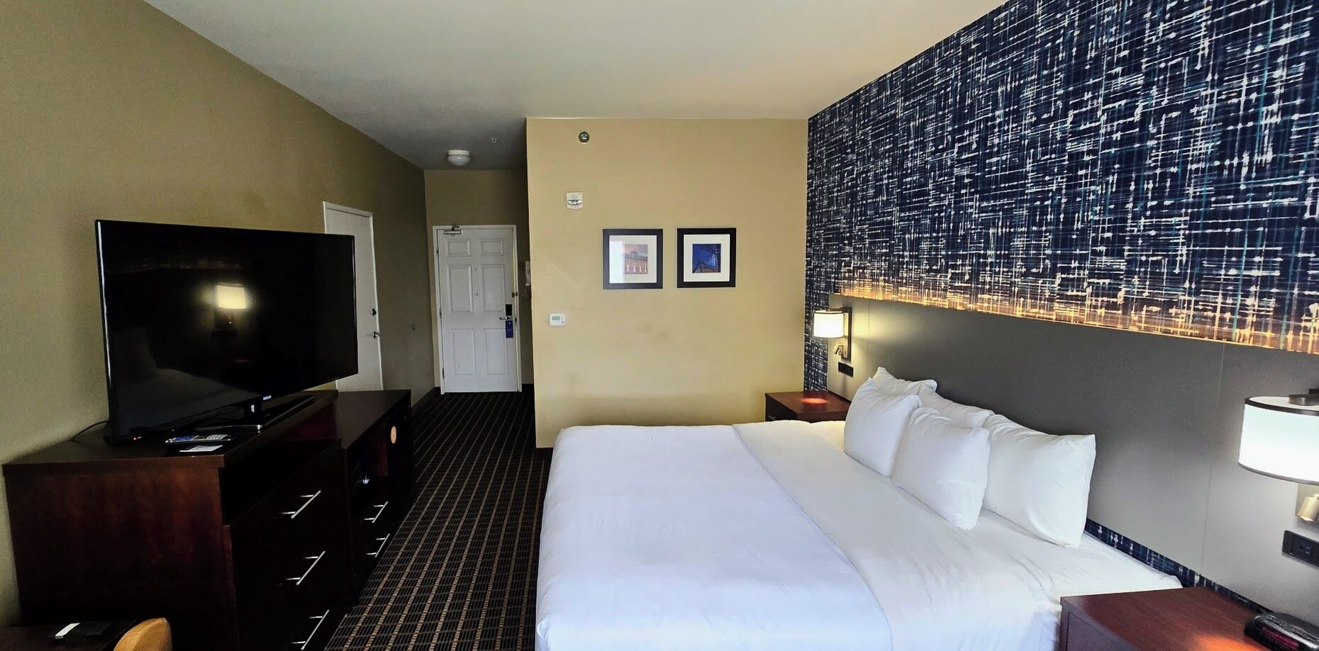 A hotel room with a bed, a TV, a wall with a patterned design, and two framed pictures on the wall.