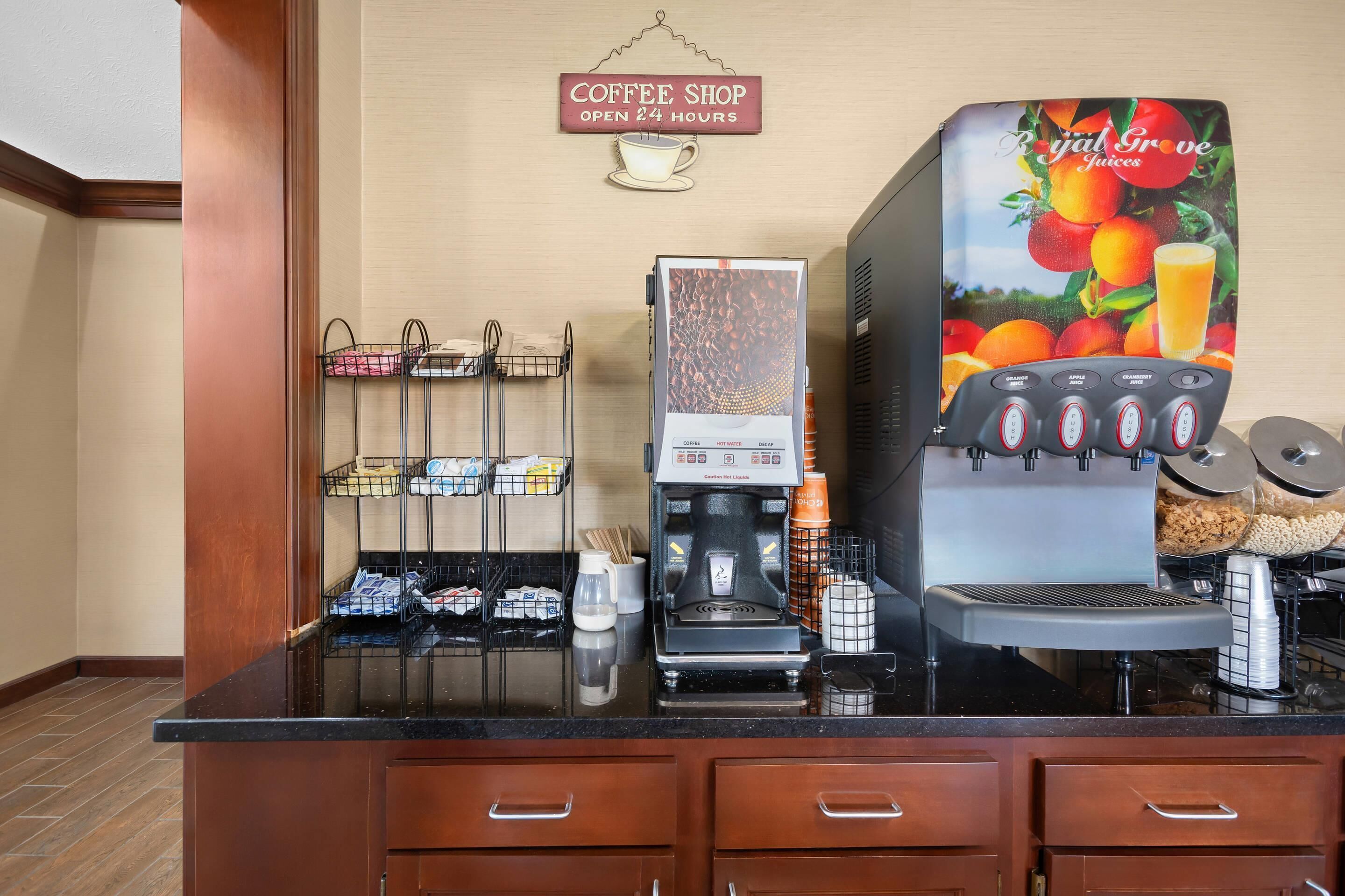 Bancada de café com máquina de café, dispensador de suco e placa indicando funcionamento 24 horas.