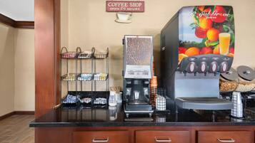 Bancada de café com máquina de café, dispensador de suco e placa indicando funcionamento 24 horas.