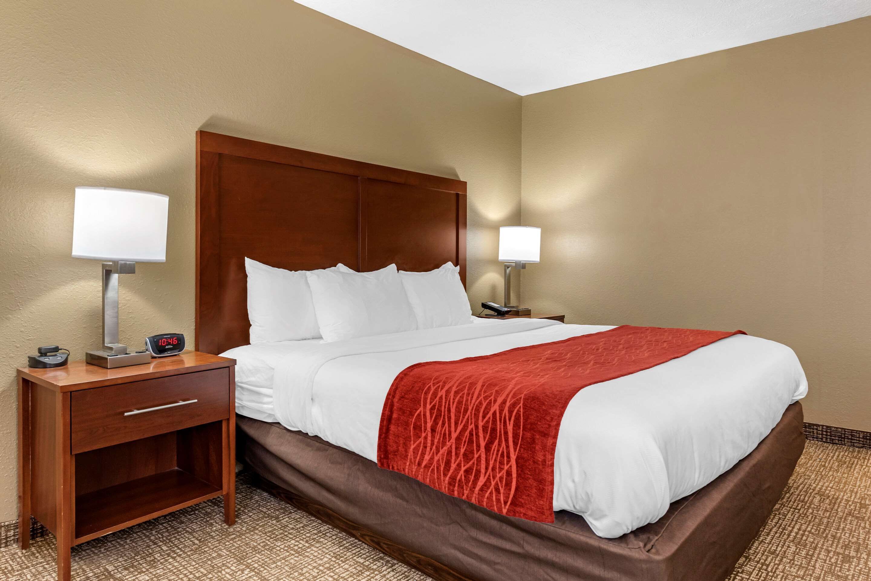 A hotel room with a large bed, two bedside tables with lamps, a nightstand with a drawer, and a red throw blanket.