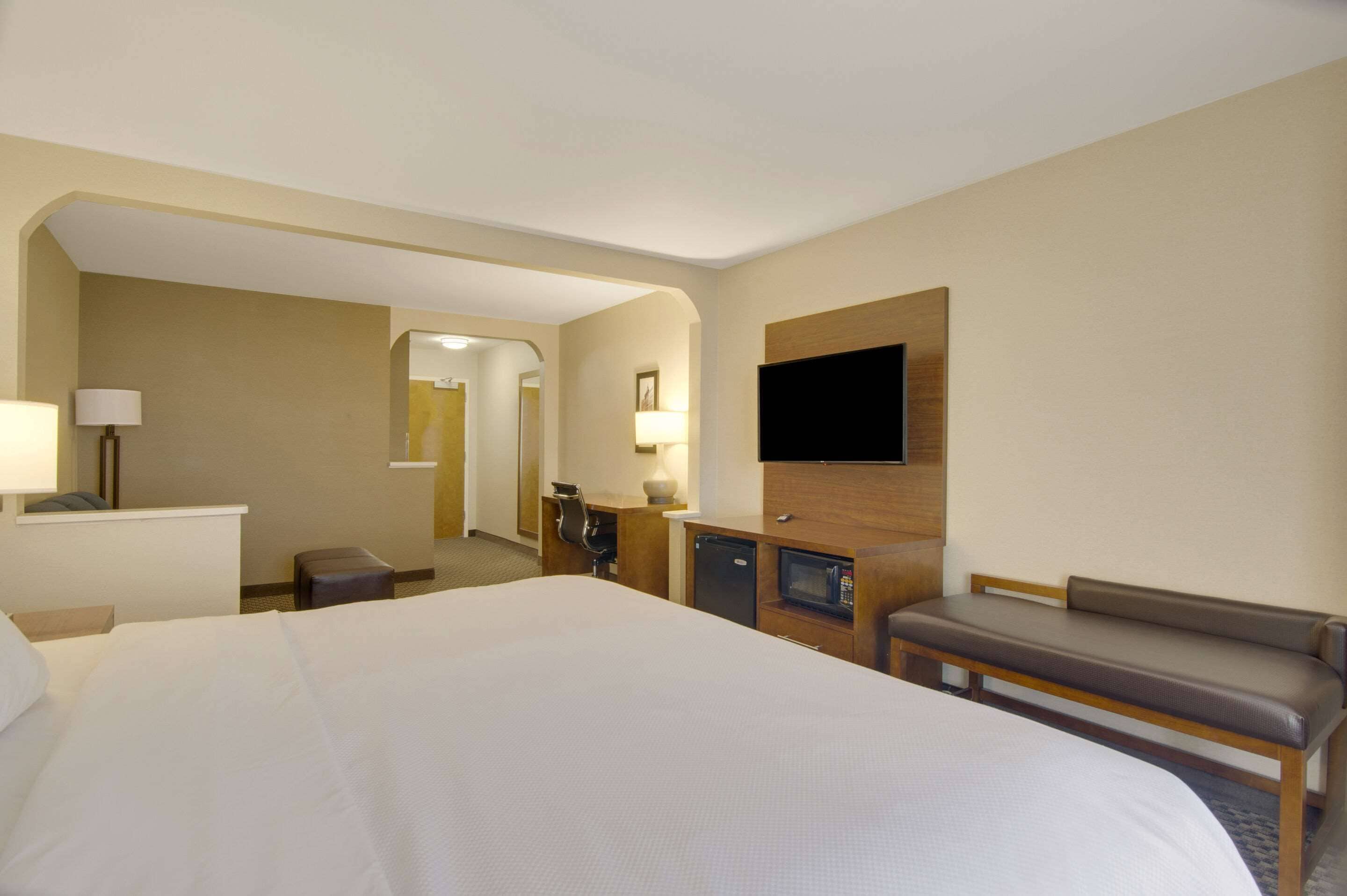 A hotel room with a large bed, a desk with a lamp, a television mounted on the wall, and a bench.