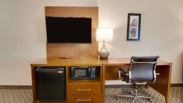 Una habitación de hotel con un escritorio de madera, un televisor de pantalla plana, microondas, refrigerador, lámpara y un cuadro en la pared.