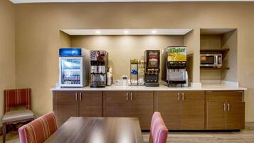 Una kitchenette de hotel con cafetera, microondas y refrigerador. Hay una mesa de comedor con sillas y un banco.