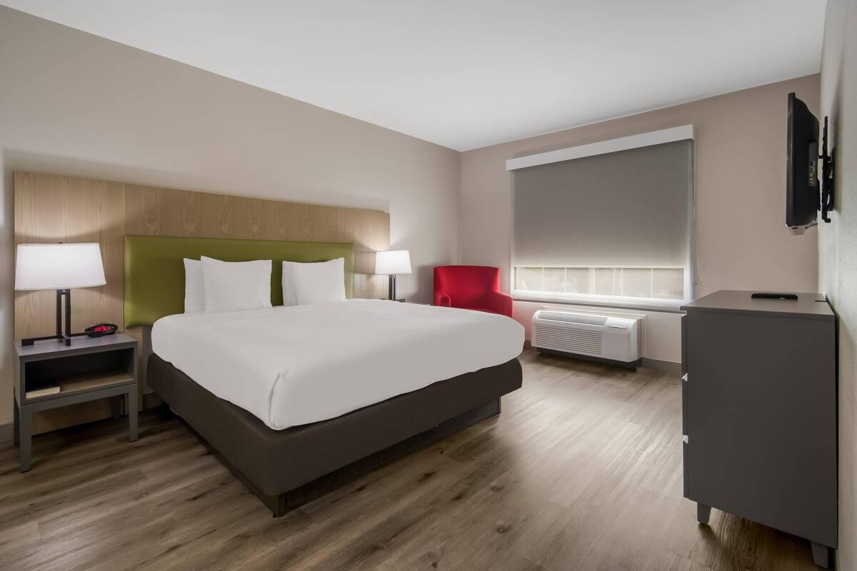 Ein Hotelzimmer mit Bett, Nachttisch, einem roten Sessel, Fernseher und einem Fenster mit Jalousien.