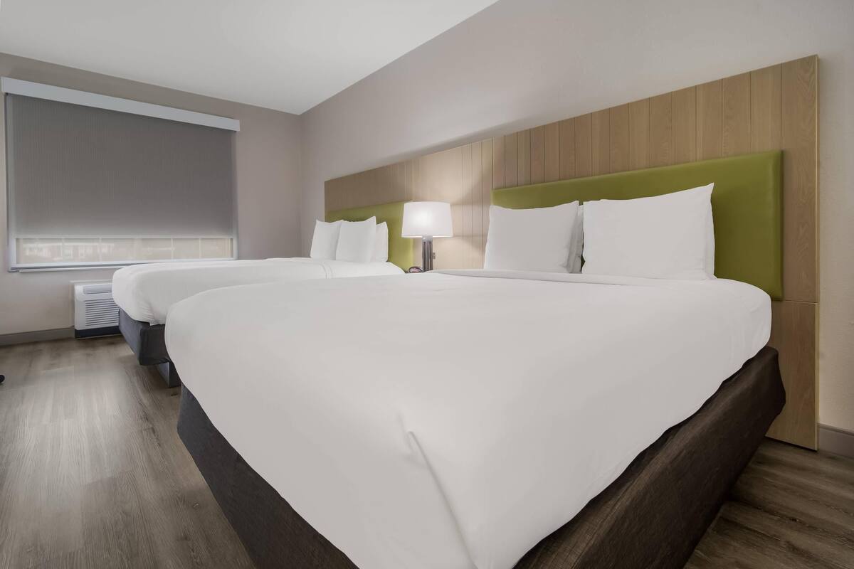 Ein Hotelzimmer mit zwei Betten, einem großen Fenster mit Jalousien, einer Wand-Klimaanlage und einer Nachttischlampe.