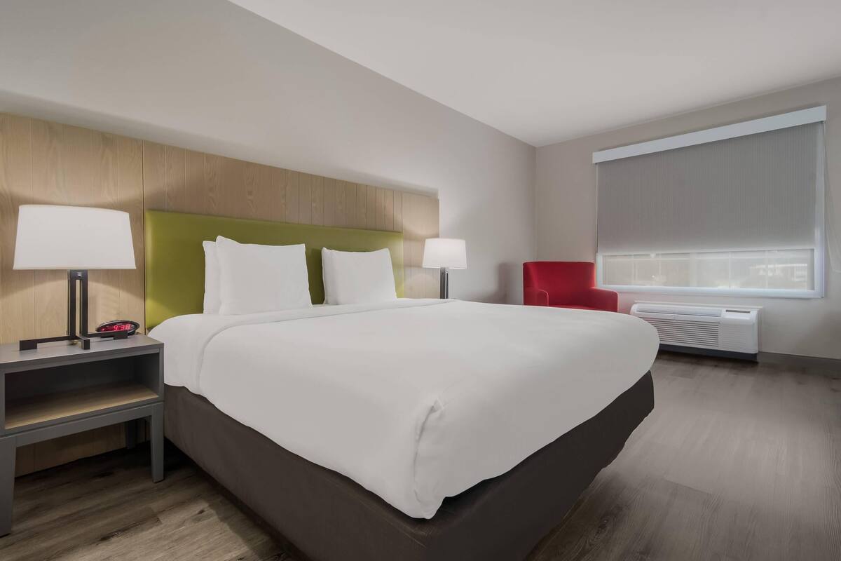 Ein Hotelzimmer mit einem großen Bett, zwei Nachttischen mit Lampen, einem roten Sessel und einem Fenster mit Jalousien.