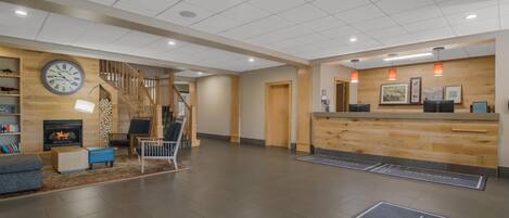 A modern hotel lobby with a reception desk, a clock, a fireplace, and a staircase.