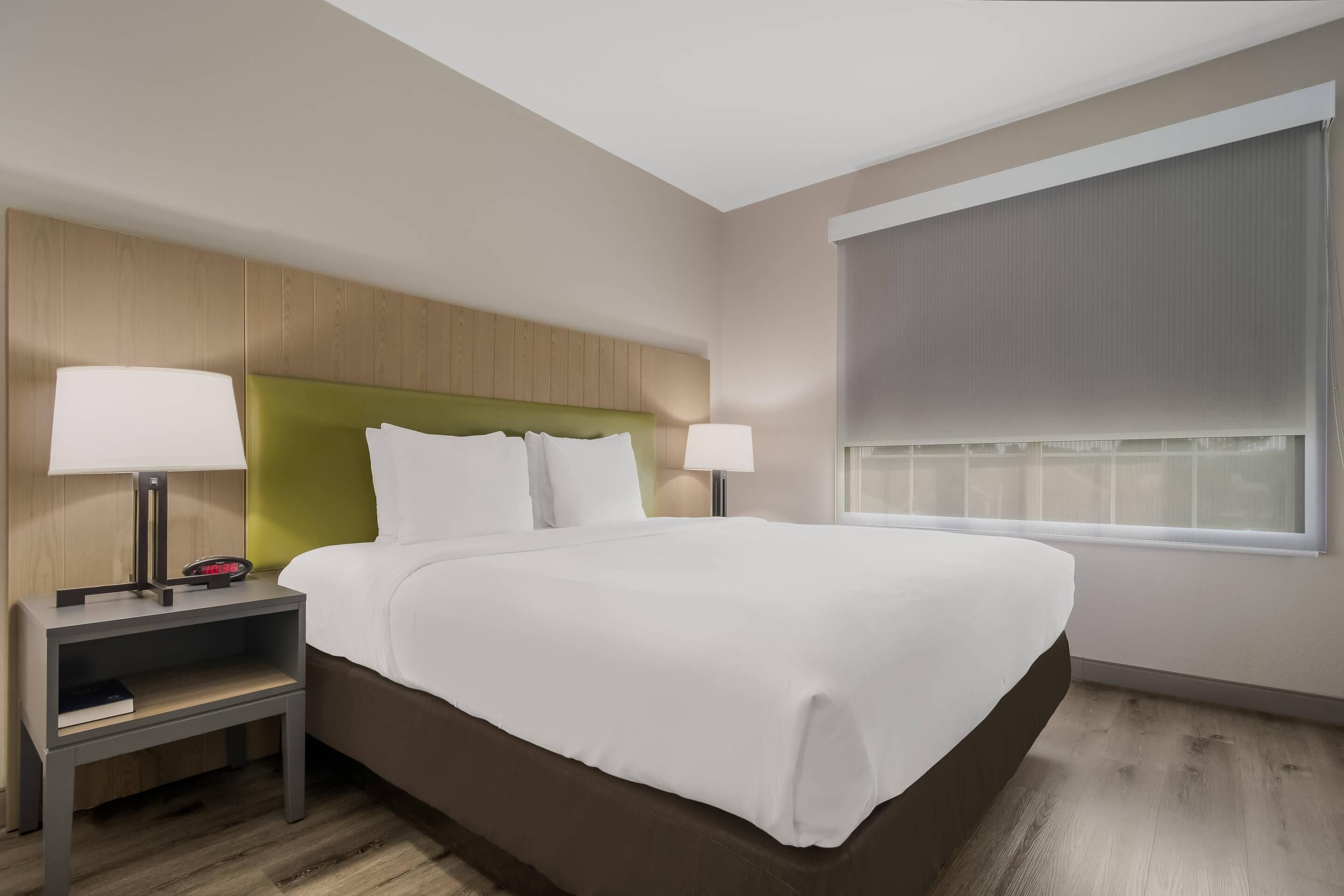 Een moderne hotelkamer met een groot bed, twee nachtkastjes met lampen, een groen hoofdeinde en een groot raam met jaloezieën.