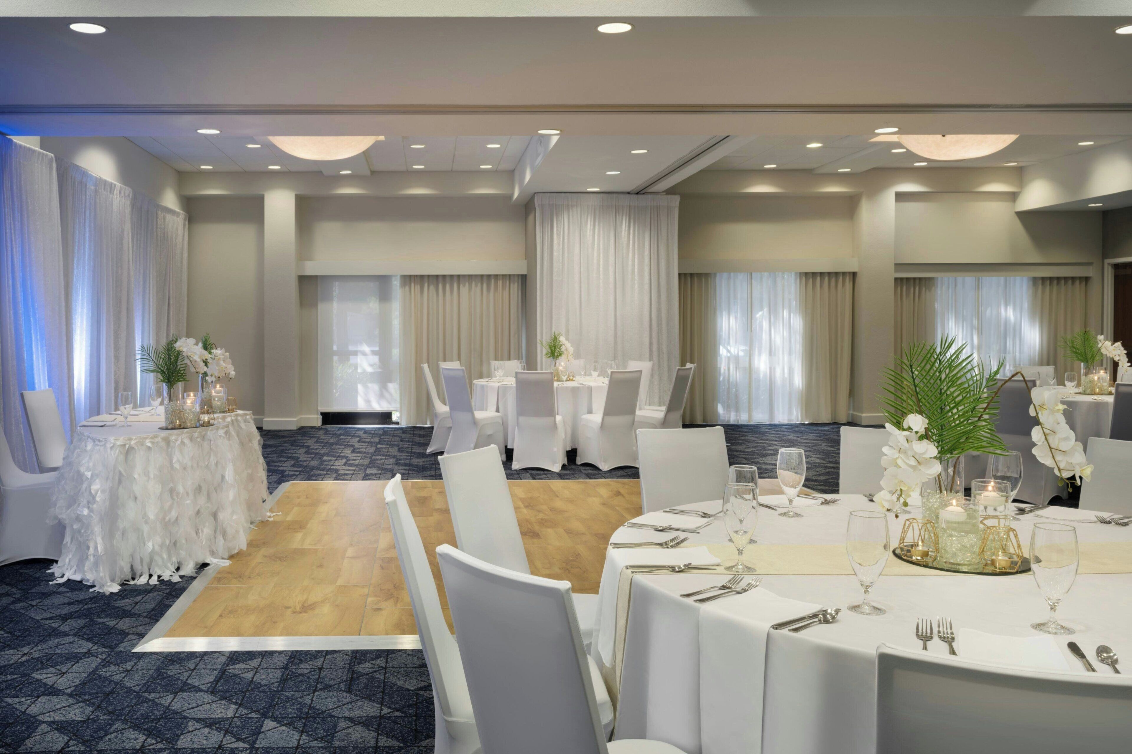 Un salón de banquetes con mesas redondas, sillas blancas y centros de mesa florales.