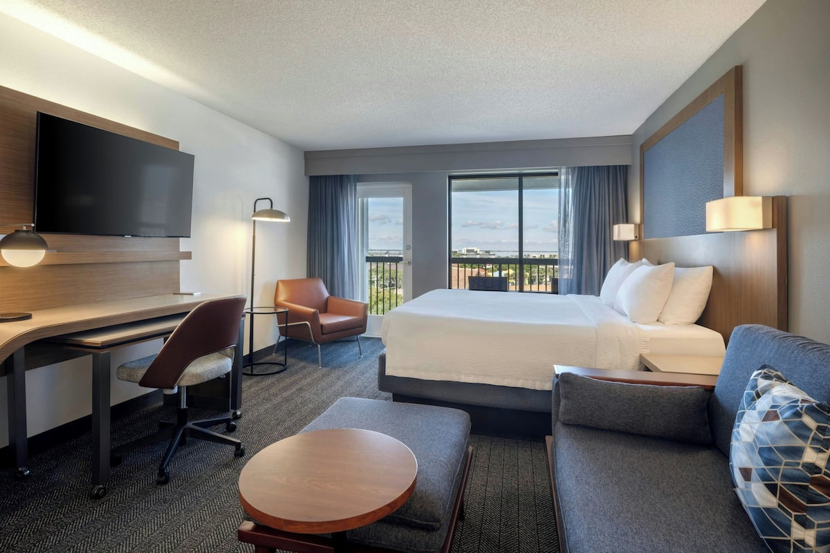 Habitación de hotel con cama, escritorio, sillón, televisor y balcón con vistas.