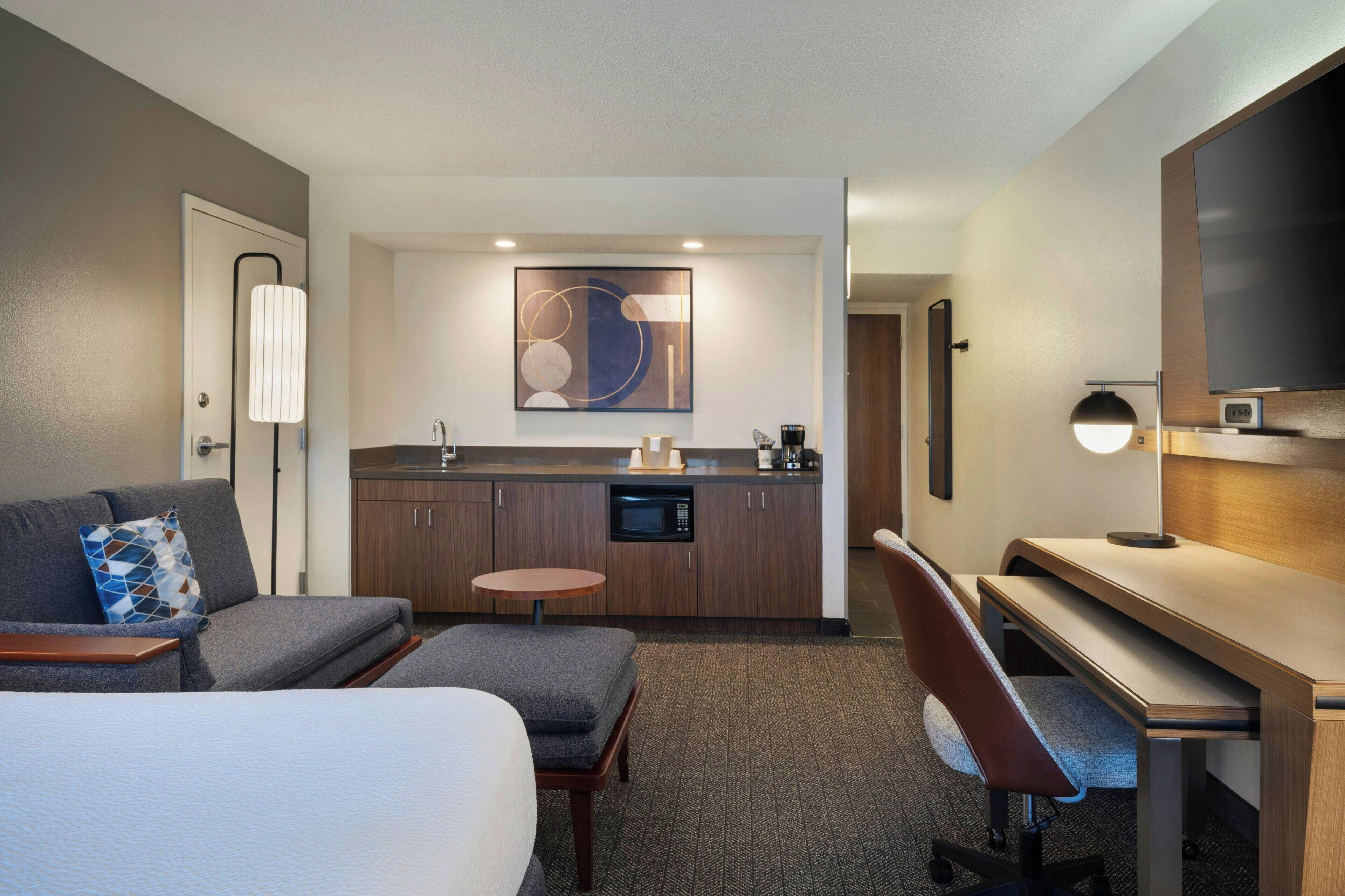 A hotel room with a bed, a sofa, a desk, a chair, a small table, a microwave, a coffee maker, and a TV.