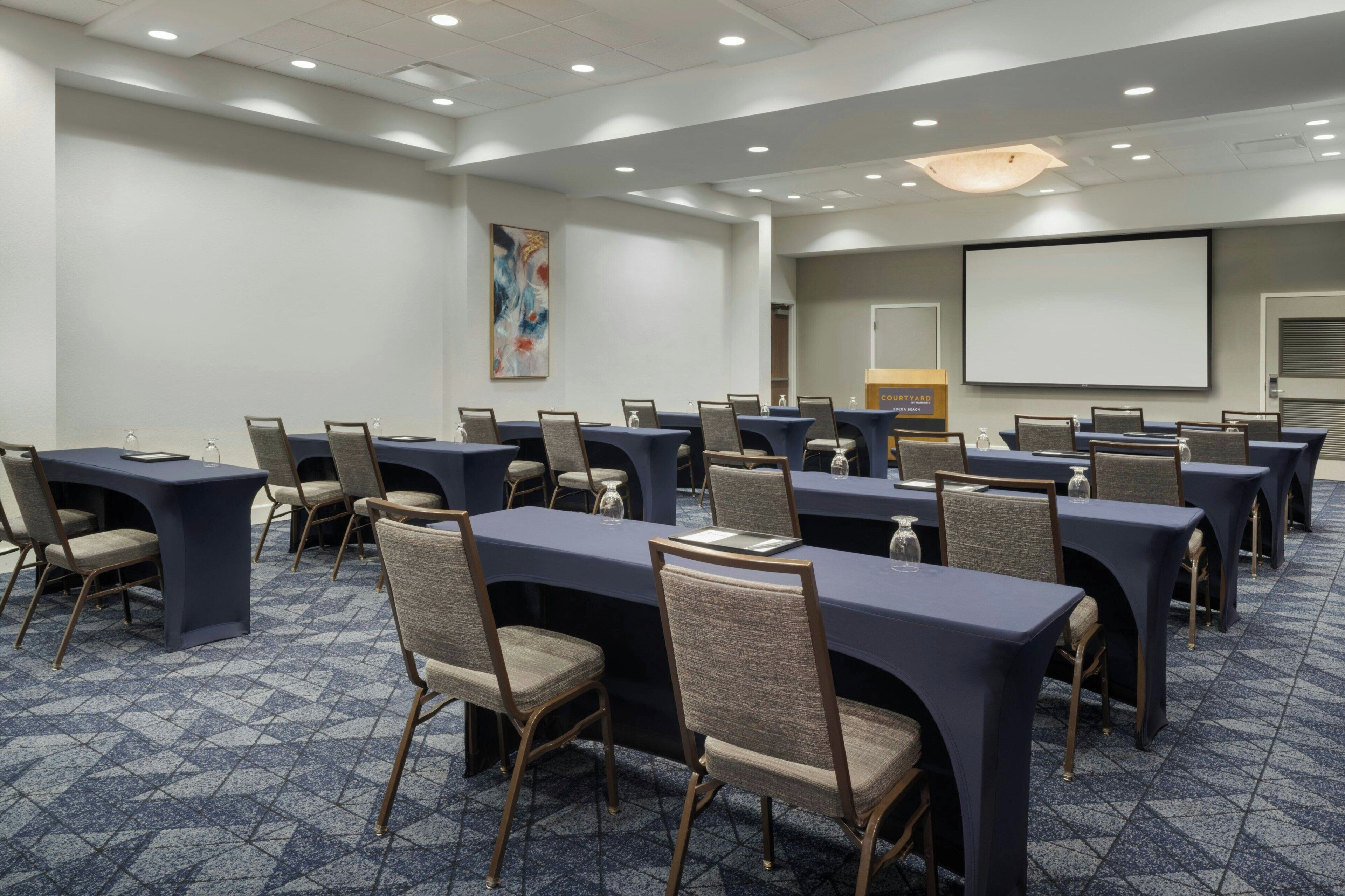 A conference room with multiple tables and chairs, a projector screen, and a large painting on the wall.