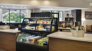 Zona de recepción de hotel con un expositor de bebidas, una máquina de café y un amplio ventanal con vistas a zonas verdes.