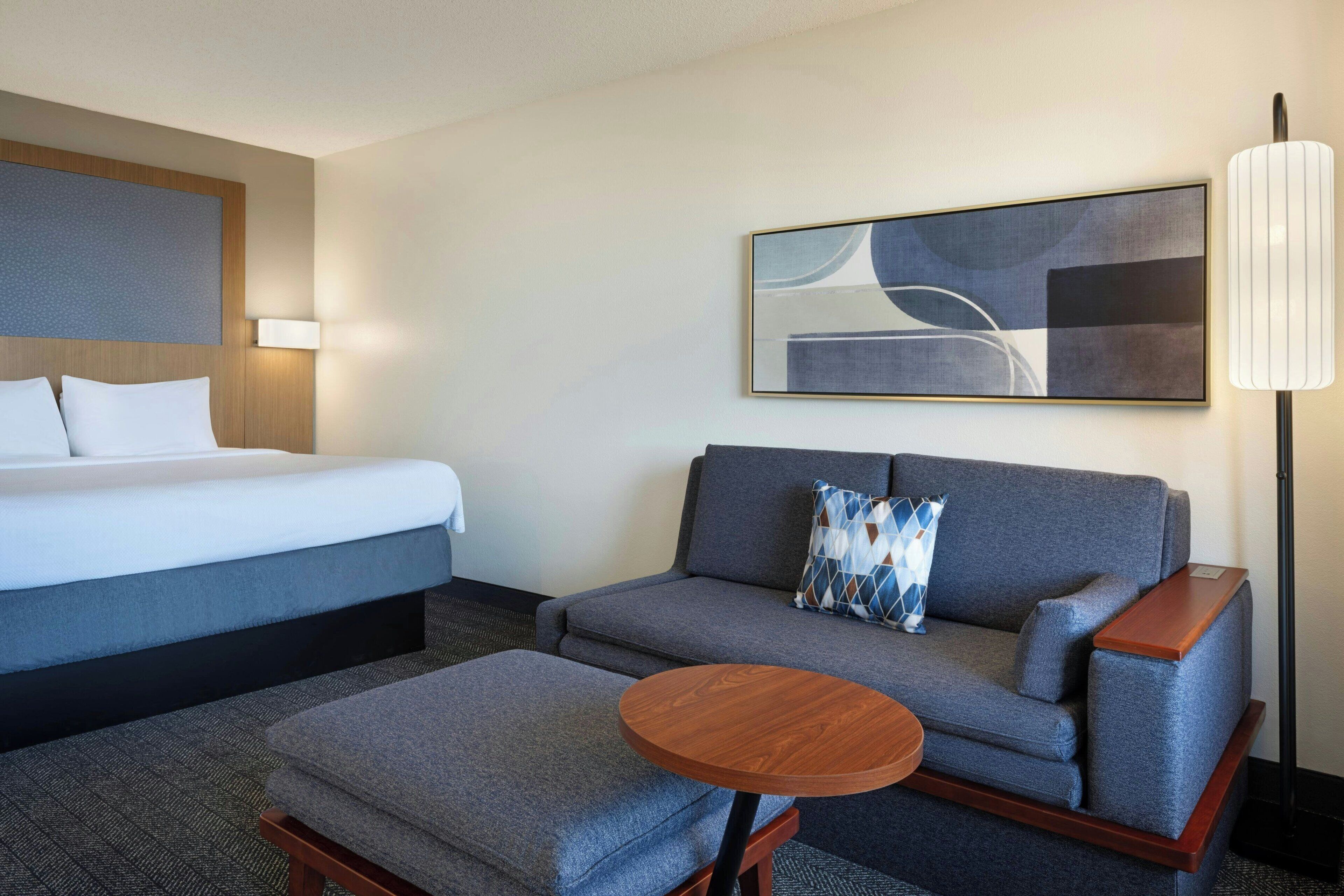 Ein Hotelzimmer mit einem Bett, einem grauen Sofa mit passendem Hocker, einem kleinen Holztisch und einer Stehlampe.
