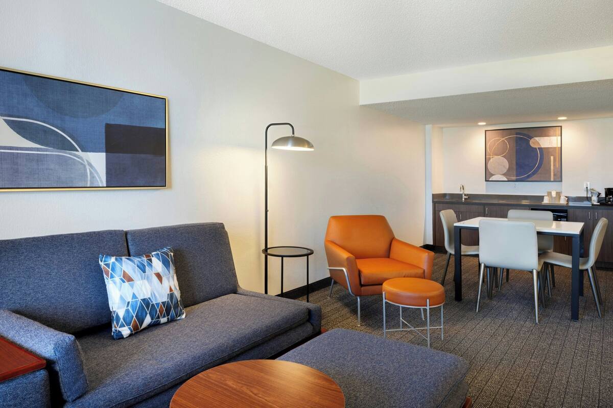 Habitación de hotel moderna con un sofá gris, un sillón naranja, una mesa de comedor con sillas, una mesita redonda pequeña y una lámpara de pie.