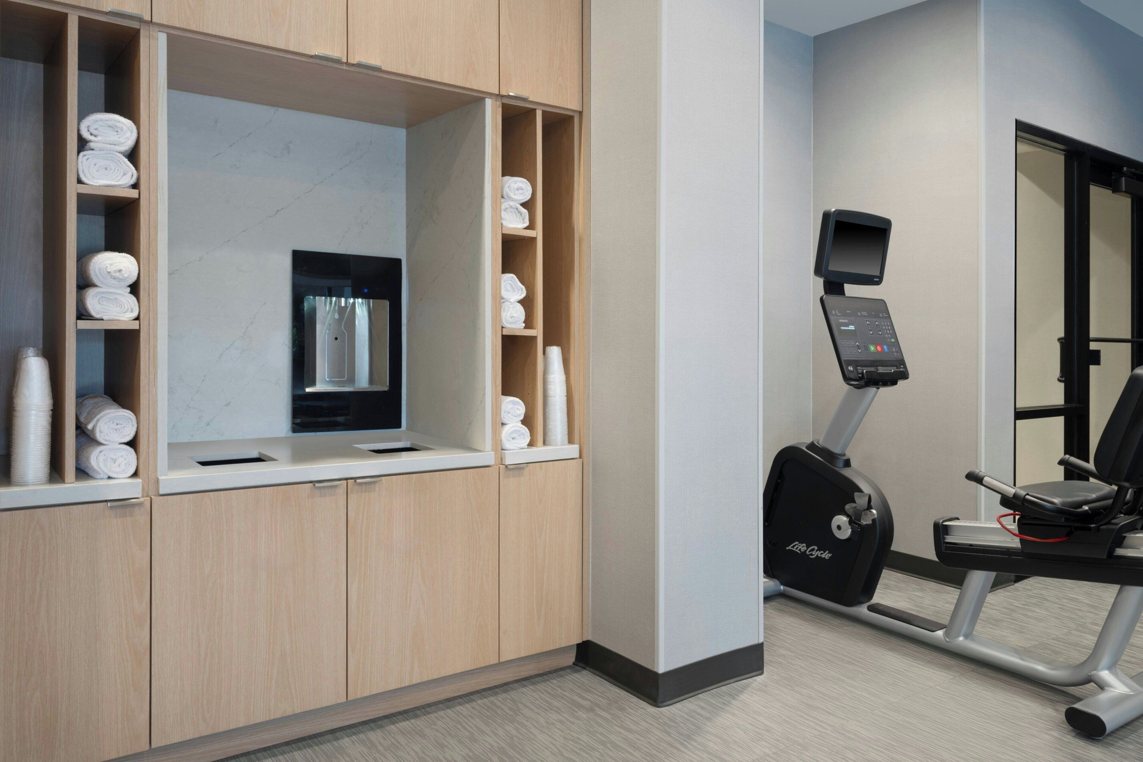 Habitación de hotel con un armario de madera, toallas, dispensador de agua y una máquina elíptica Life Fitness.