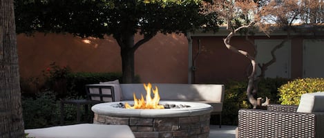 A circular stone fire pit with a fire, surrounded by outdoor furniture including a sofa and chairs, set against a backdrop of trees and a building wall.