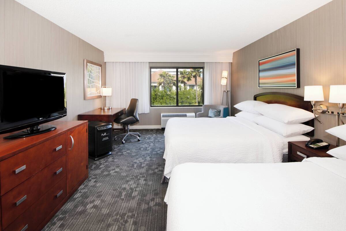 Habitación de hotel con televisión, escritorio, dos camas, una silla y ventana con vista a palmeras.