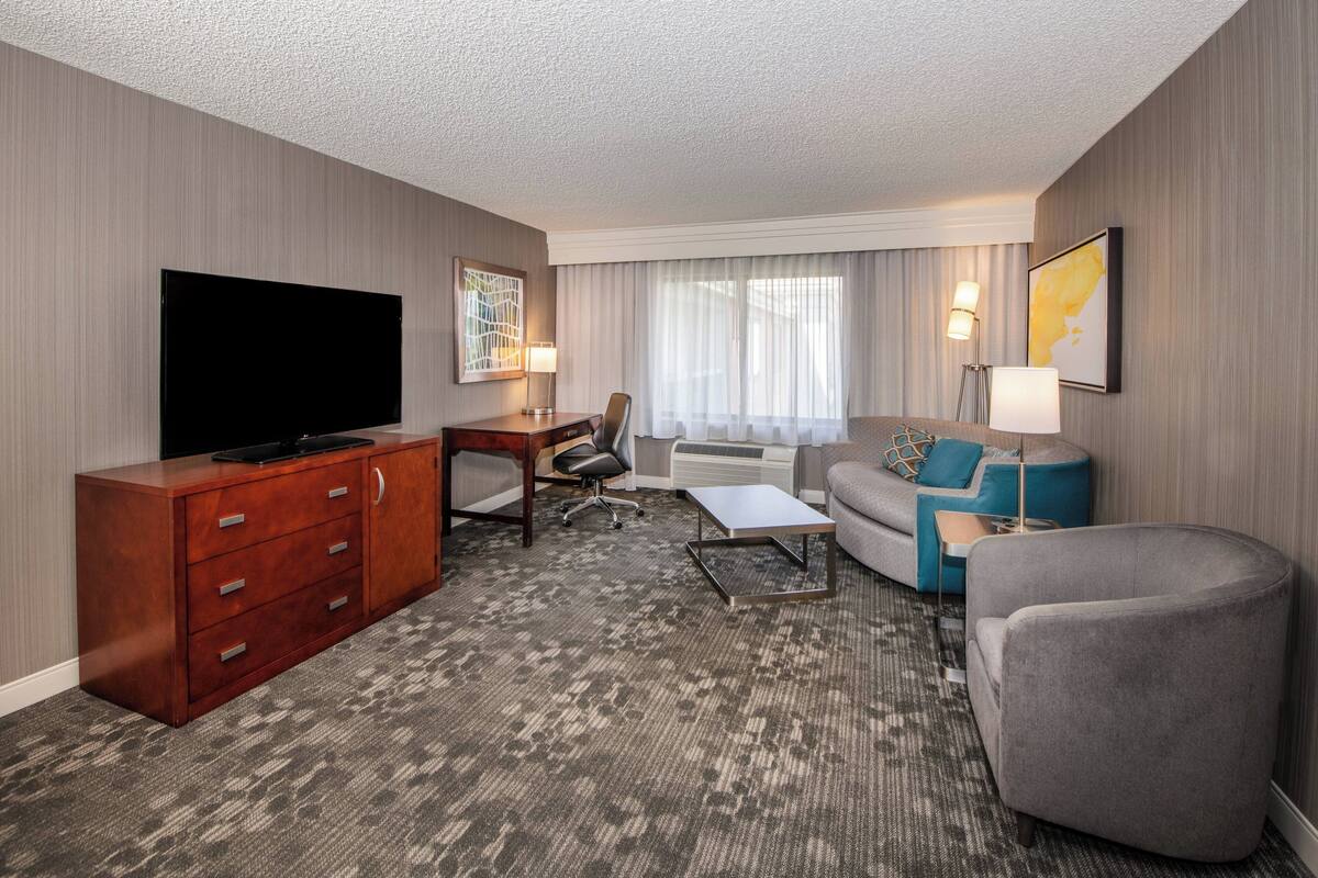 Habitación de hotel con televisor de pantalla plana, cómoda de madera, sofá gris, sillón gris, mesita de centro pequeña, escritorio con silla y ventana grande con cortinas.
