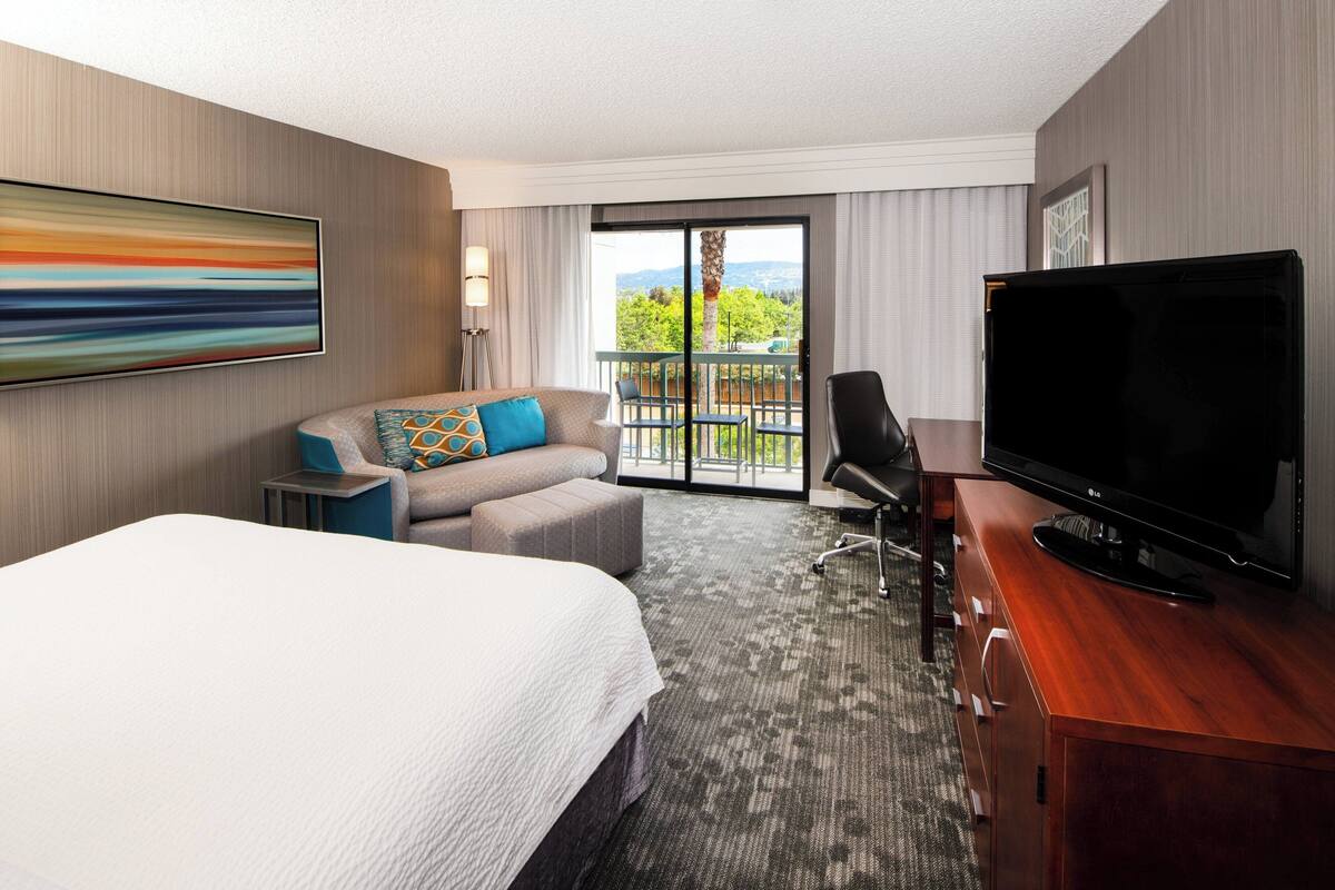 Una habitación de hotel con una cama grande, un sofá con cojines de colores, un televisor de pantalla plana, un aparador de madera y un balcón con vistas a zonas verdes.