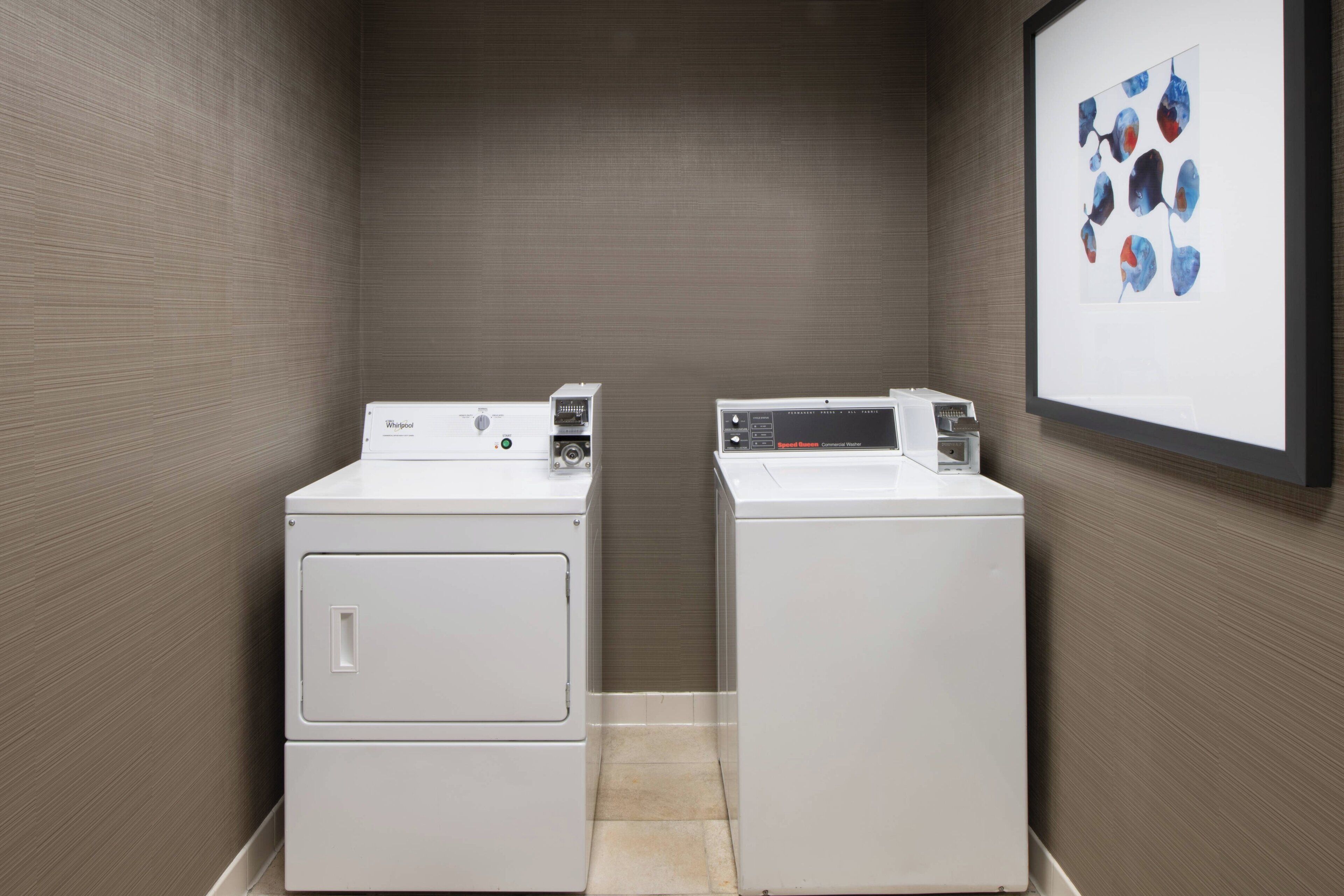A laundry room with two white front-loading washers and dryers, a tiled floor, and a framed abstract art piece on the wall.