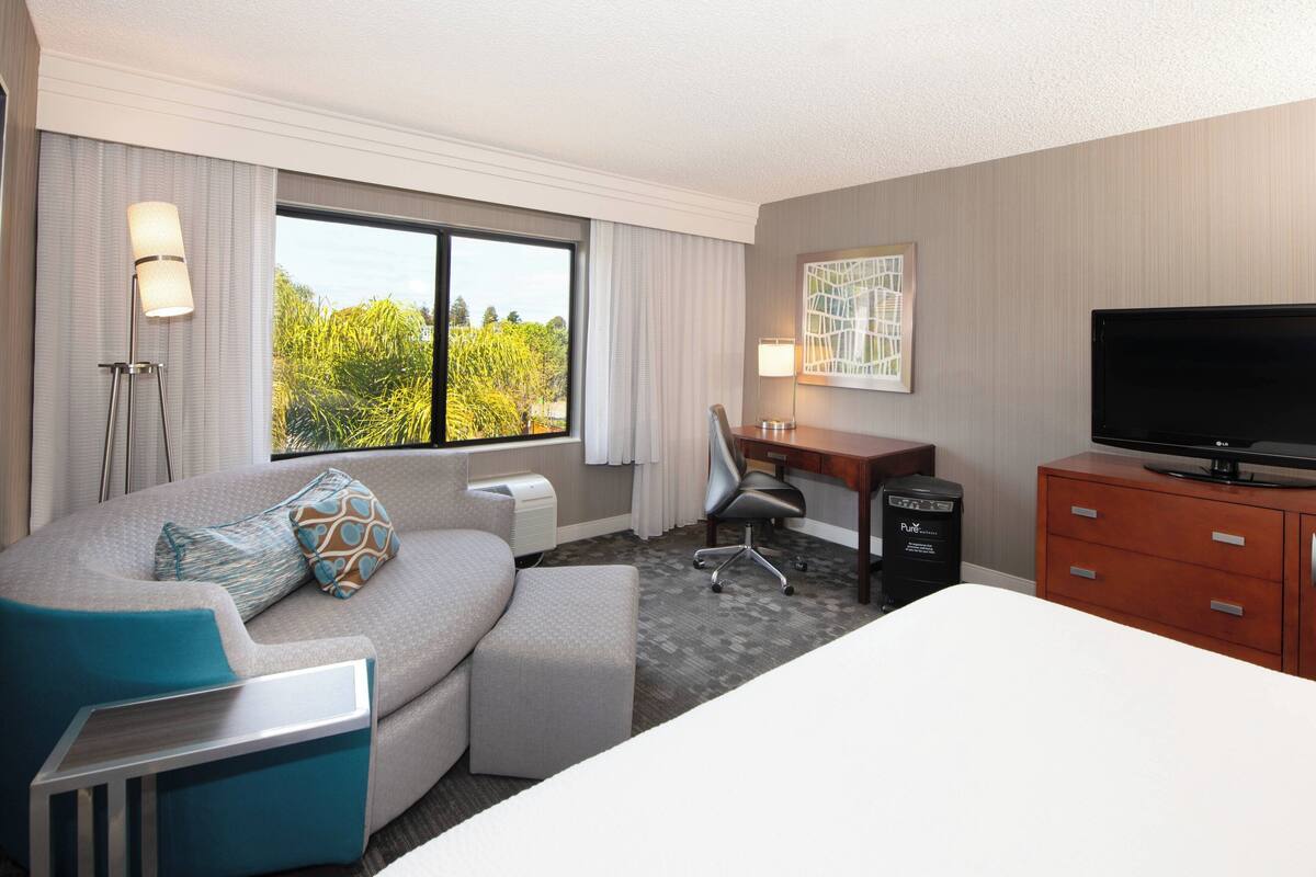 Habitación de hotel con una cama grande, un sofá con reposapiés, un escritorio con silla, un televisor y una ventana con cortinas.