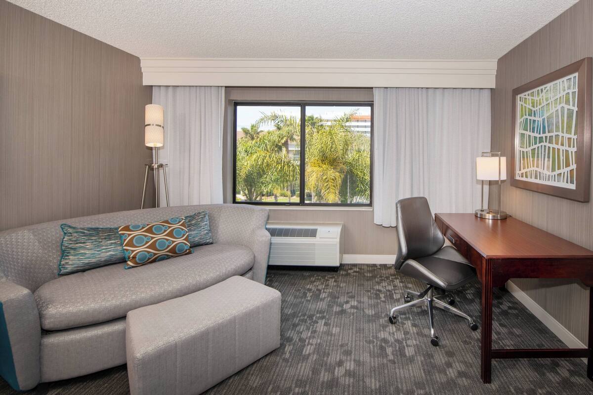Habitación de hotel con un sofá gris, un escritorio de madera, una silla y un ventanal con vistas a palmeras.