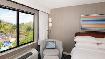 A hotel room with a large window, a chair with a pillow, a bedside table, and a bed with pillows.