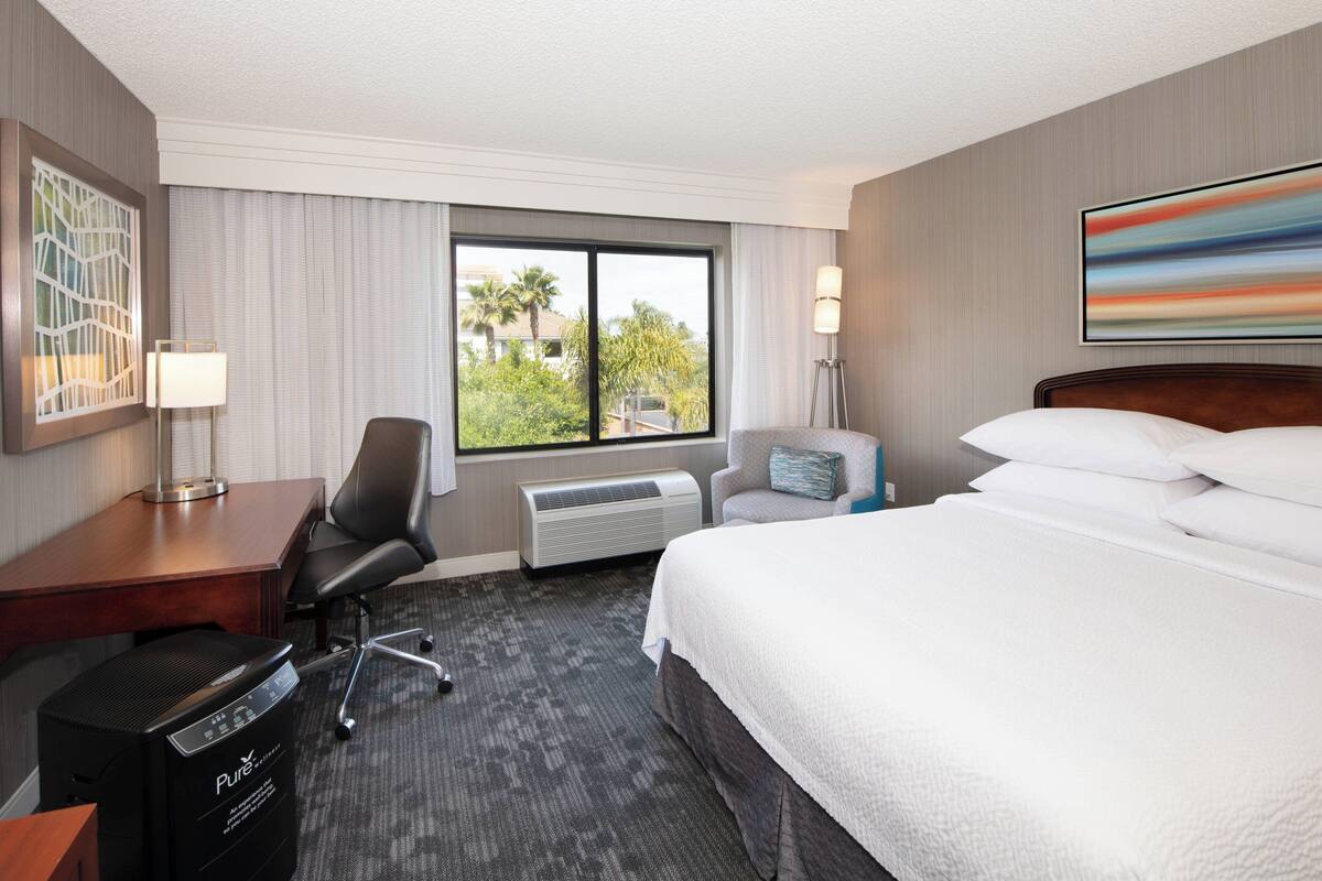 Habitación de hotel con cama, escritorio, silla, equipo de aire acondicionado y vistas a zonas verdes.